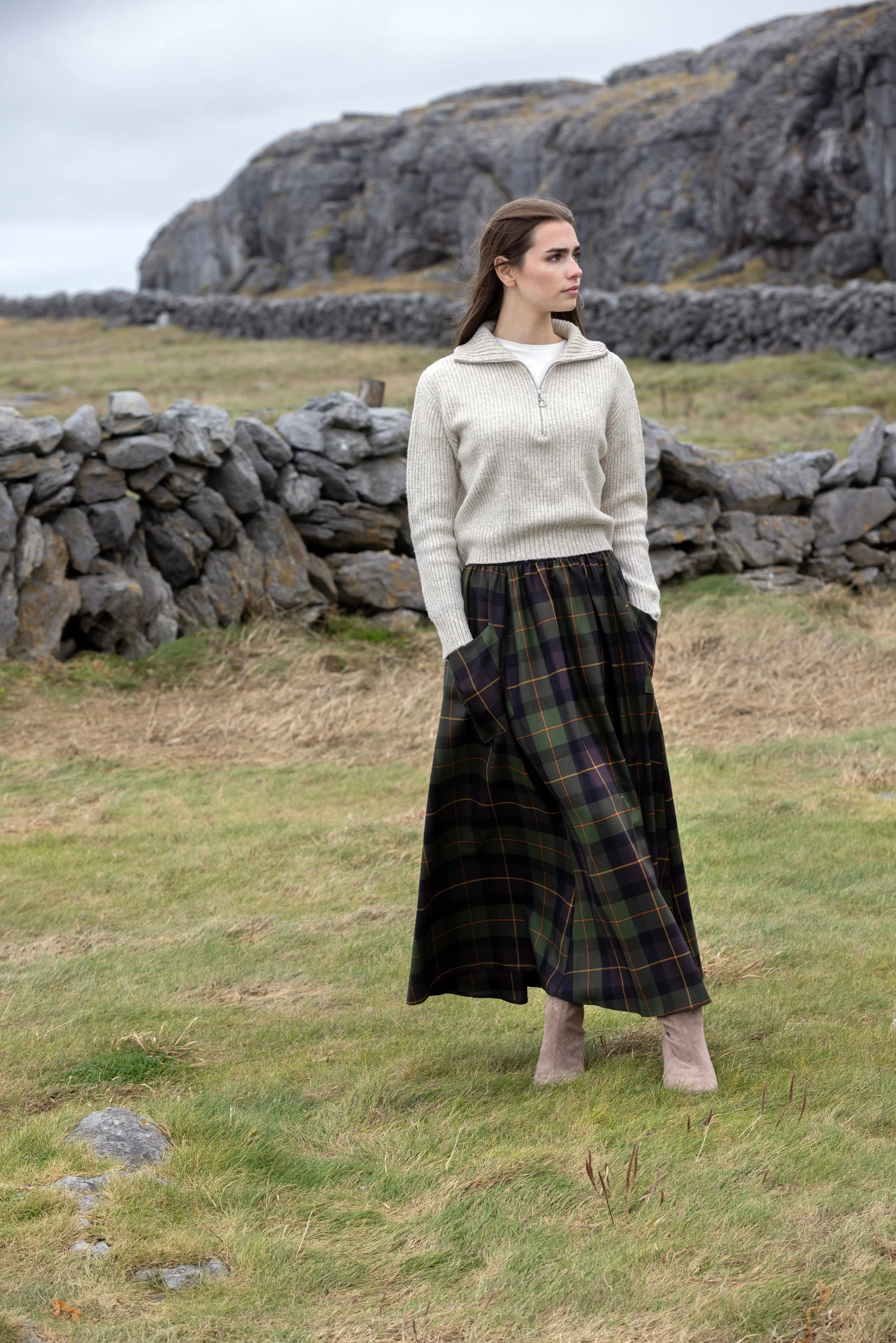 A woman standing on a grassy field with stone walls and large rocky hills in the background. She is dressed in a cream half-zip sweater, a long plaid skirt, and taupe boots, looking to her left.