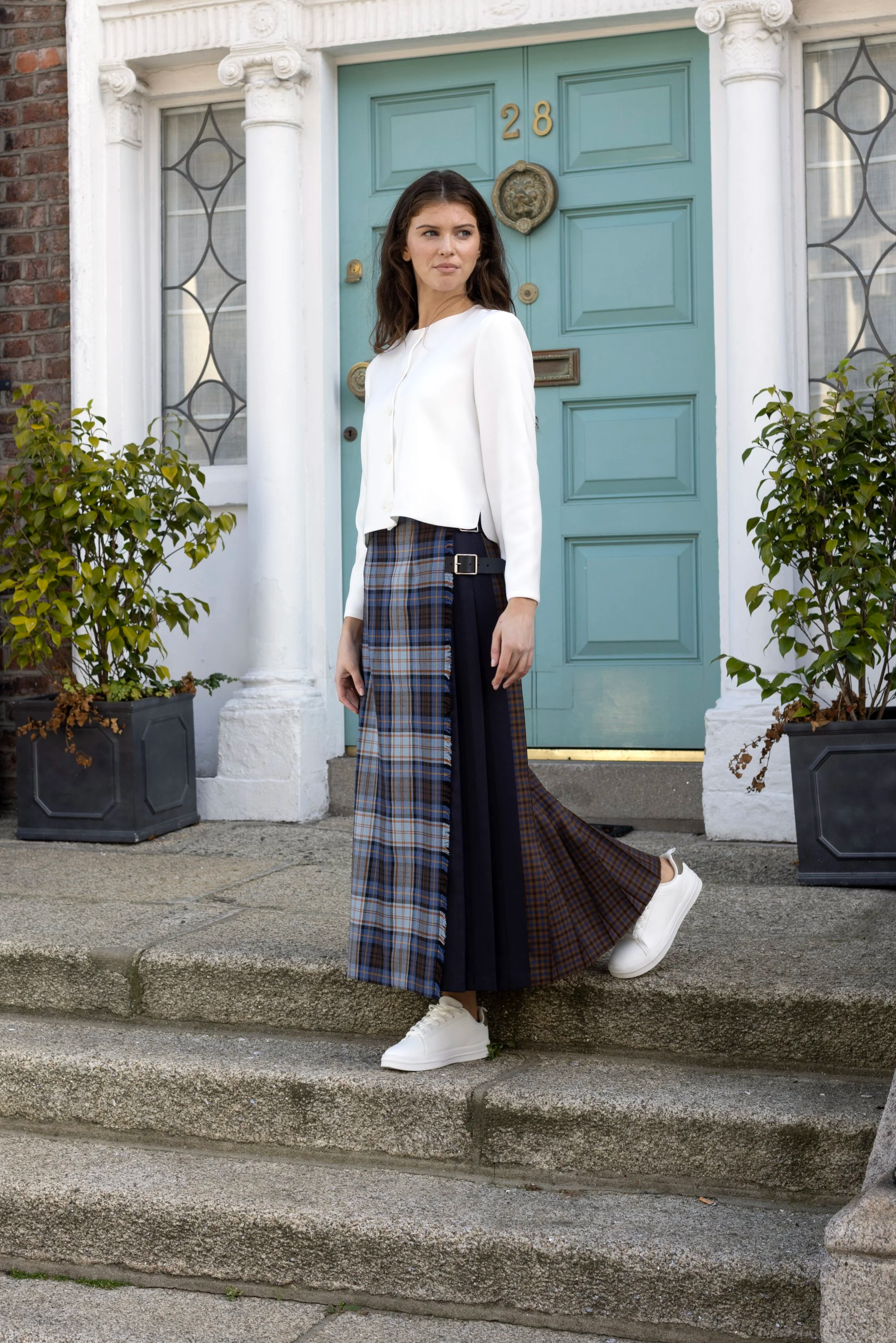 A young woman standing on the steps of a house with a bright blue door, wearing a white jacket, a layered plaid skirt, and white sneakers.