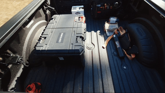 The back of a pickup truck loaded with a large black toolbox, a small white box, a portable speaker, a folded orange safety vest, and a spare tire.