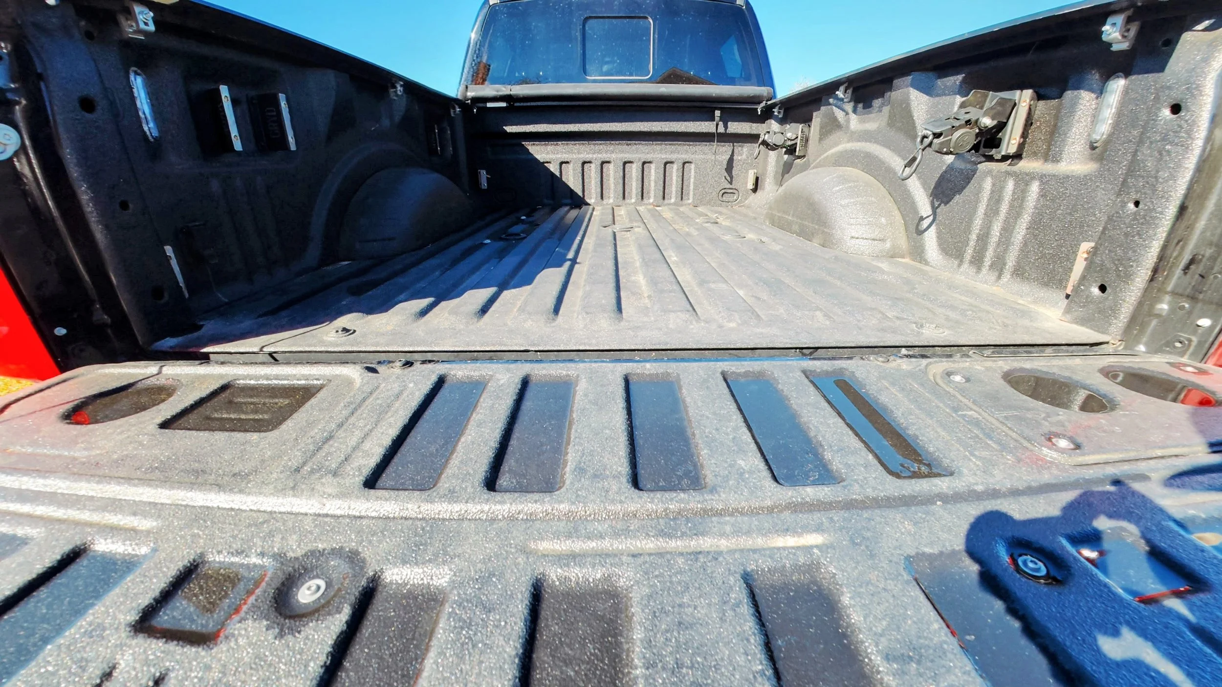 Empty truck bed with a textured surface, inside the truck bed are sidewalls with attached tools and components, with the tailgate down and a vehicle in the background under a clear sky.