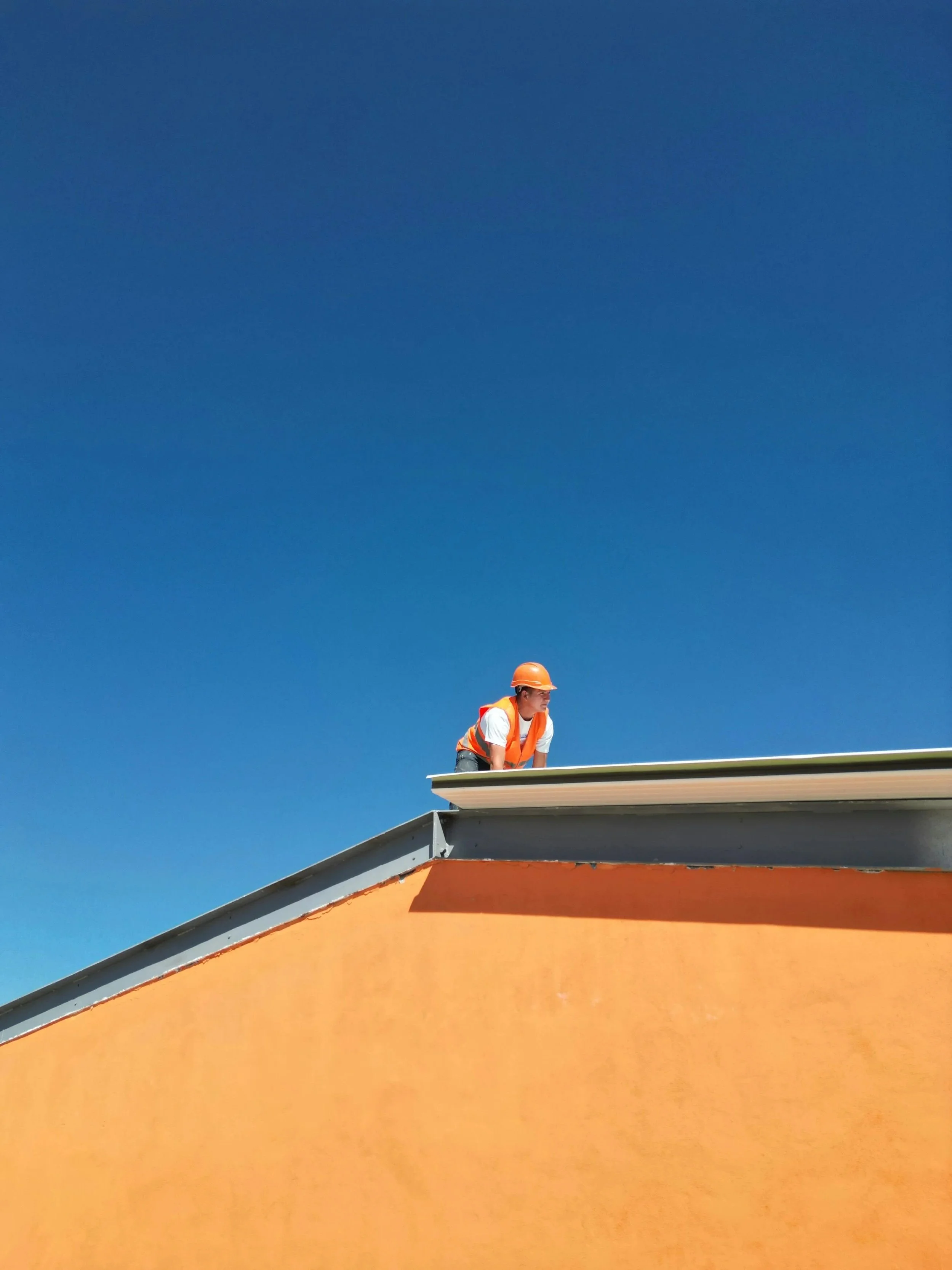 A construction worker wearing an orange helmet and safety vest crouching on the edge of a building roof, with a clear blue sky in the background.