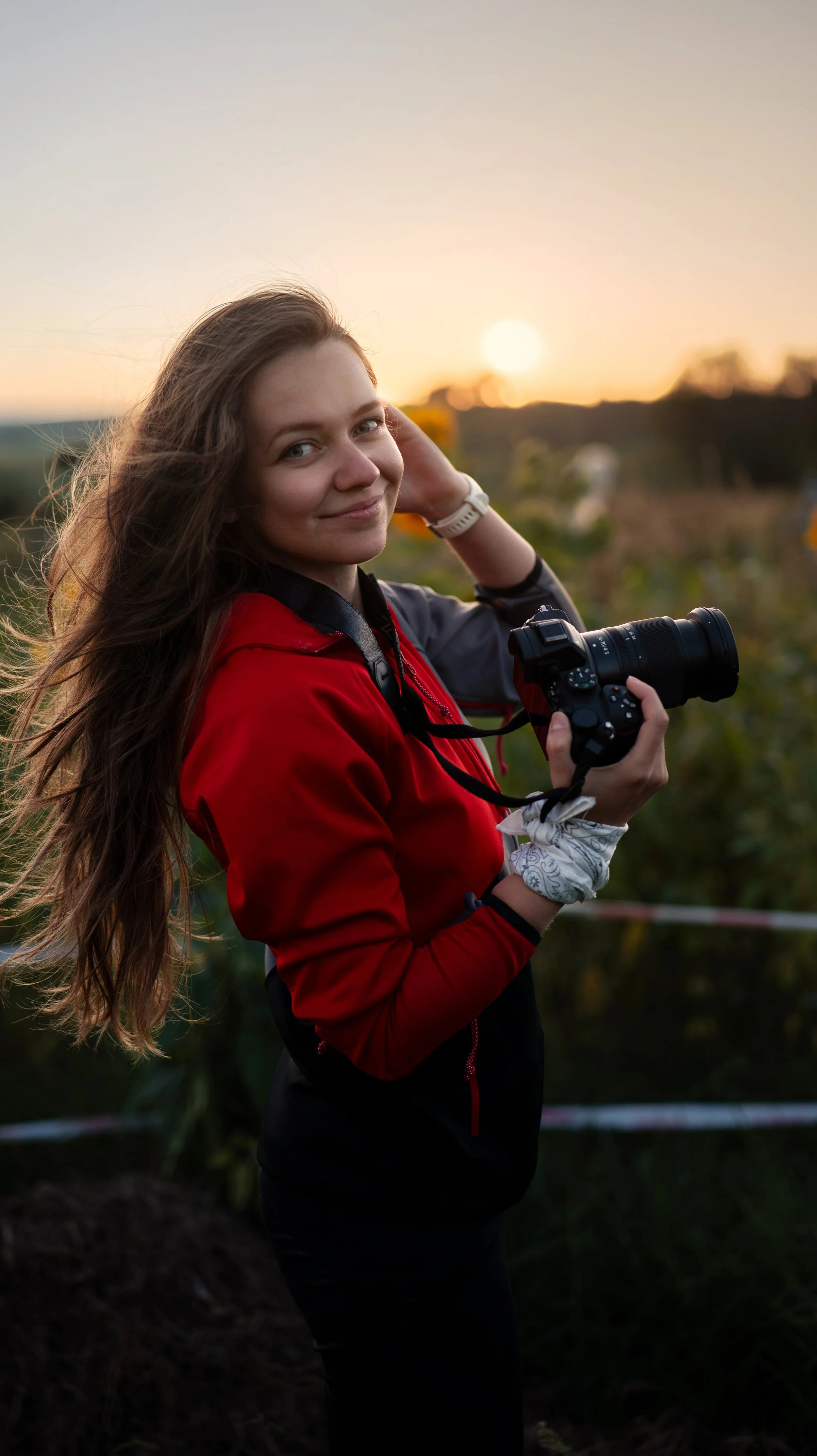 A young woman in a red and black jacket holding a camera, smiling at the camera, with a sunset in the background. Andrea Cabajova, Girl with Bandana.