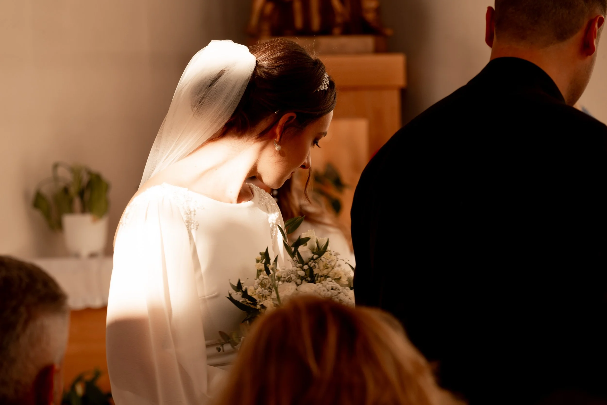 A bride in a white wedding dress with a veil and pearl earrings, holding a bouquet, during a wedding ceremony. Photographed by Andrea Cabajova, Girl with Bandana.