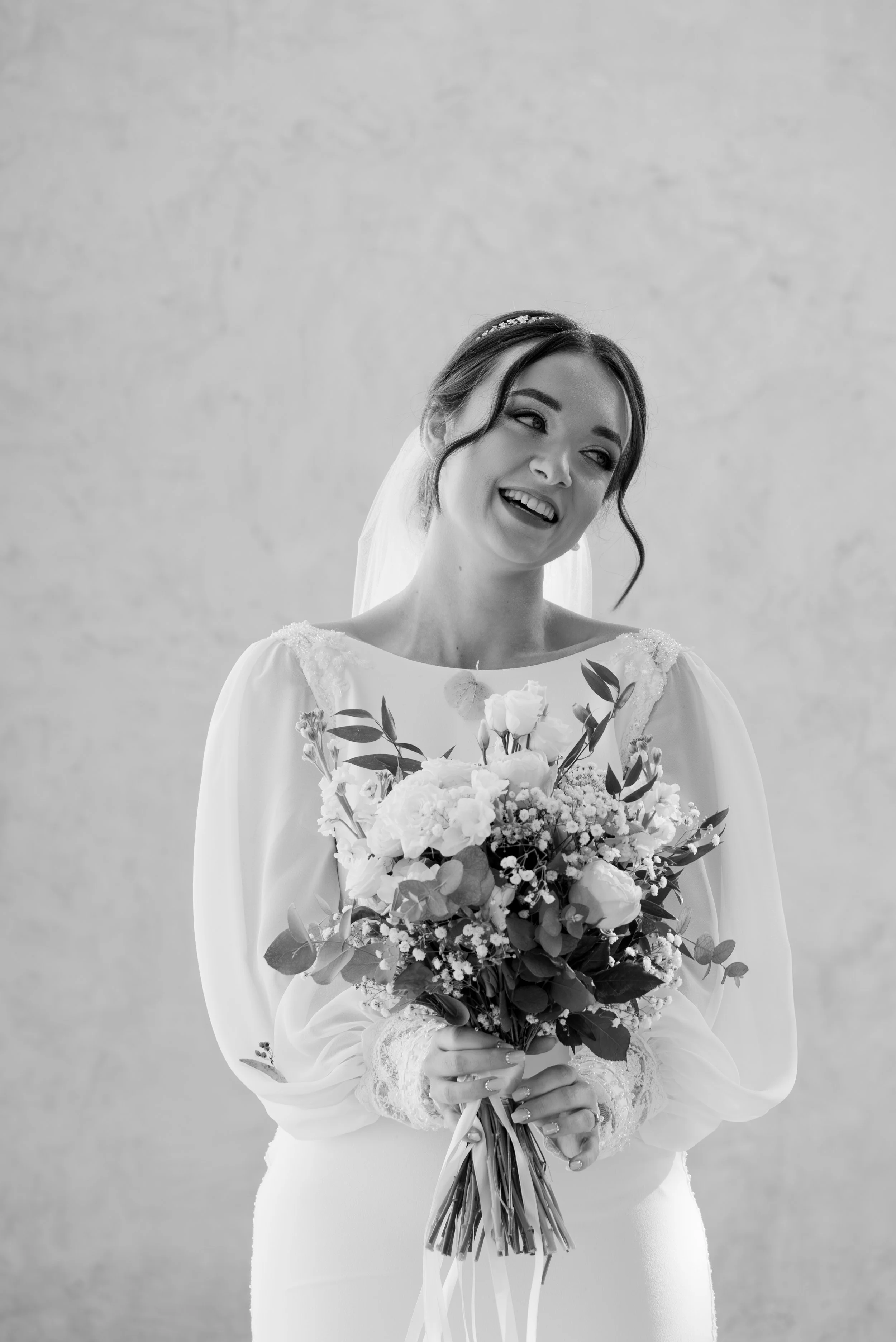 Black and white photo of a smiling bride holding a bouquet of flowers, wearing a wedding dress with long sleeves and lace details. Photographed by Andrea Cabajova, Girl with Bandana.