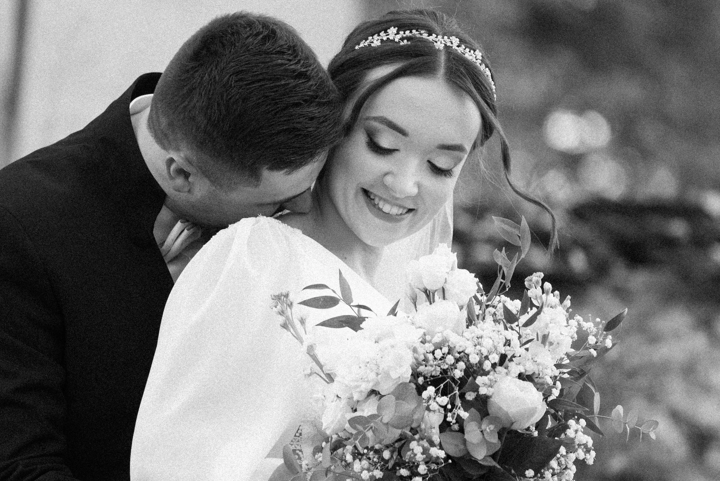 Black and white photo of a bride and groom on wedding day. The bride is smiling, holding a large bouquet, and wearing a headband. The groom is leaning in close to her, with his head near shoulder. Photographed by Andrea Cabajova, Girl with Bandana
