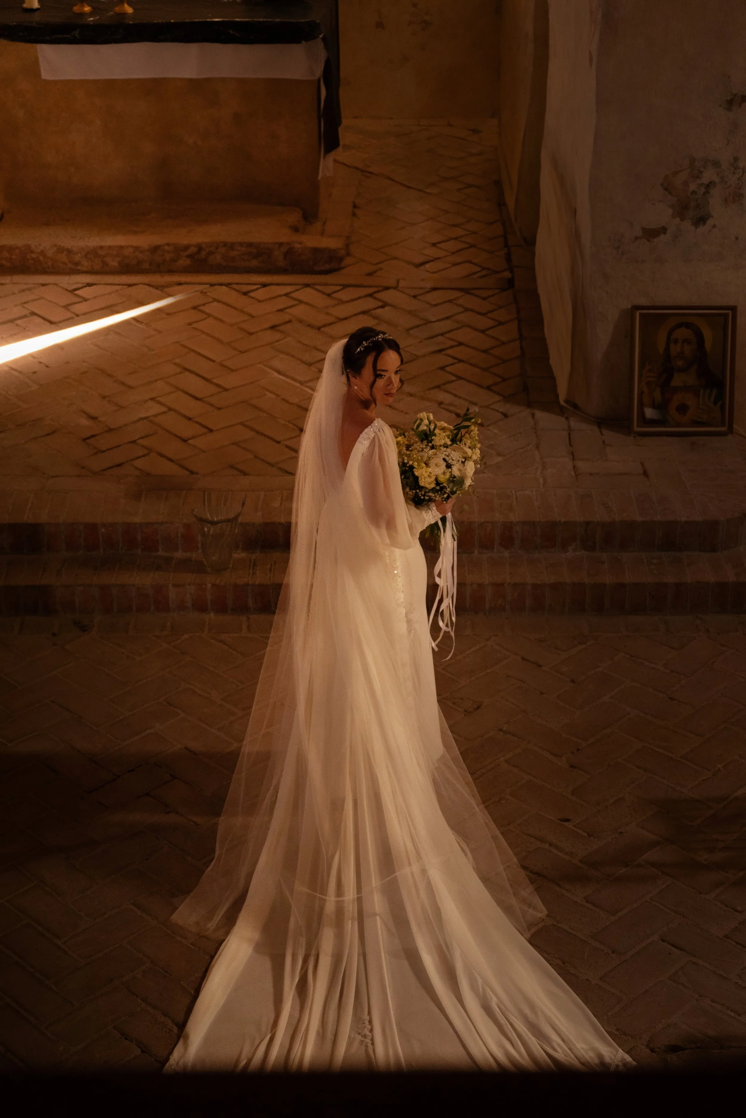 A bride in a white wedding dress with a long train, holding a bouquet of white and yellow flowers, stands inside a dimly lit church with brick flooring. An icon of Jesus is pictured on the wall to the right. Photographed by Andrea Cabajova, Girl with
