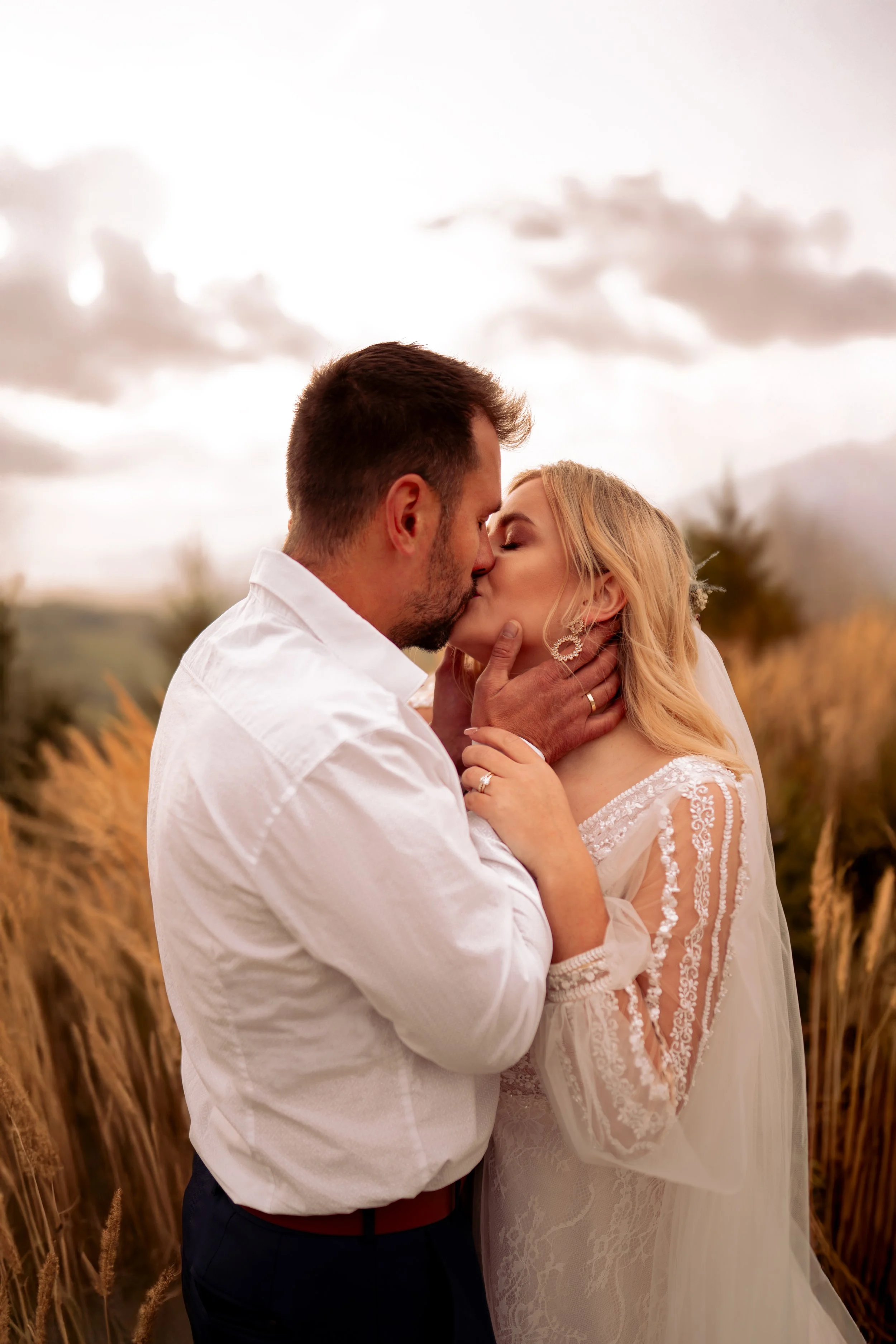 A couple sharing a kiss outdoors during sunset, with a background of tall grass and a cloudy sky. Photographed by Andrea Cabajova, Girl with Bandana.