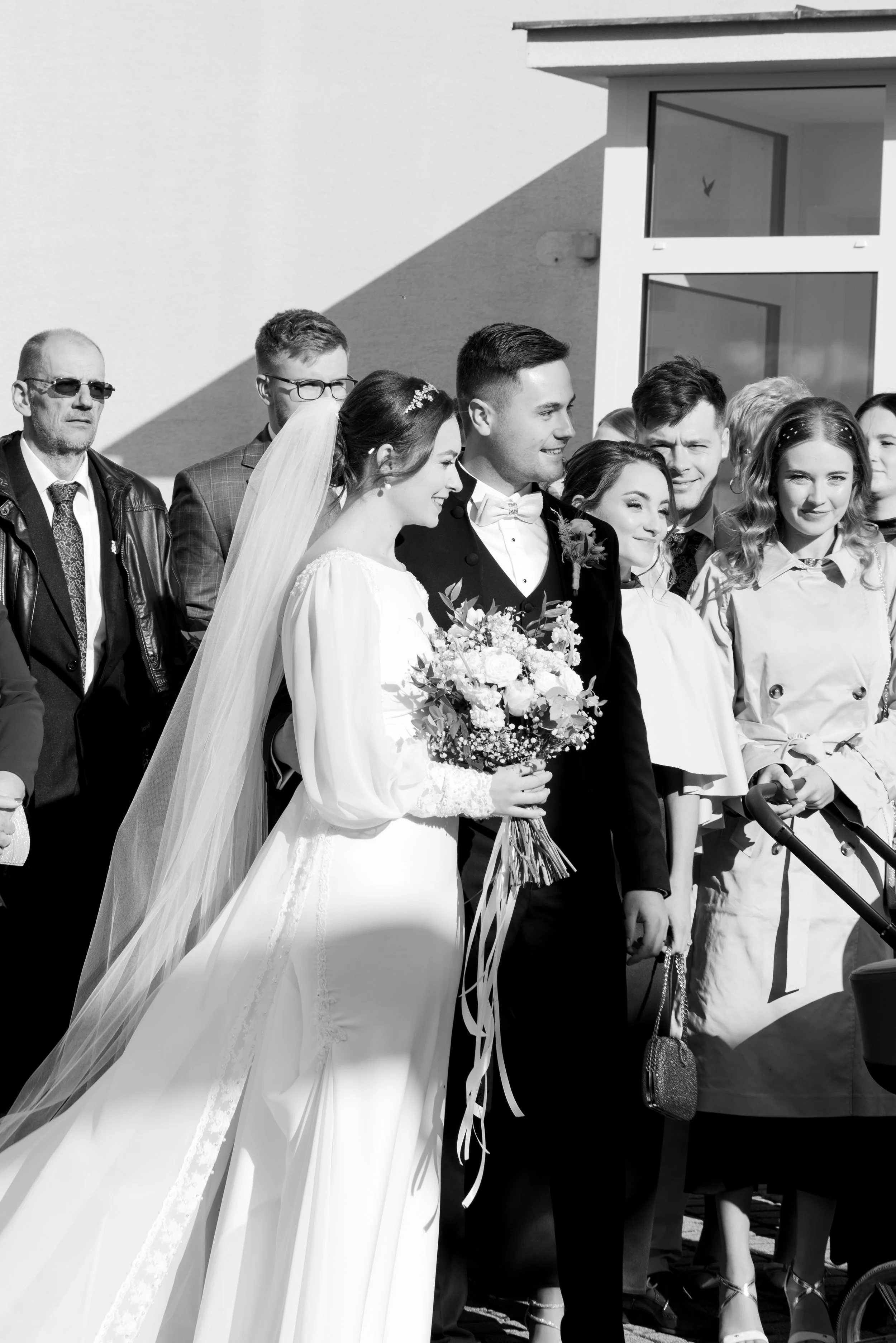 Black and white photo of a wedding crowd, including the bride in a white gown and veil holding a bouquet, and the groom in a tuxedo, standing outside near a building with large windows. Photographed by Andrea Cabajova, Girl with Bandana.