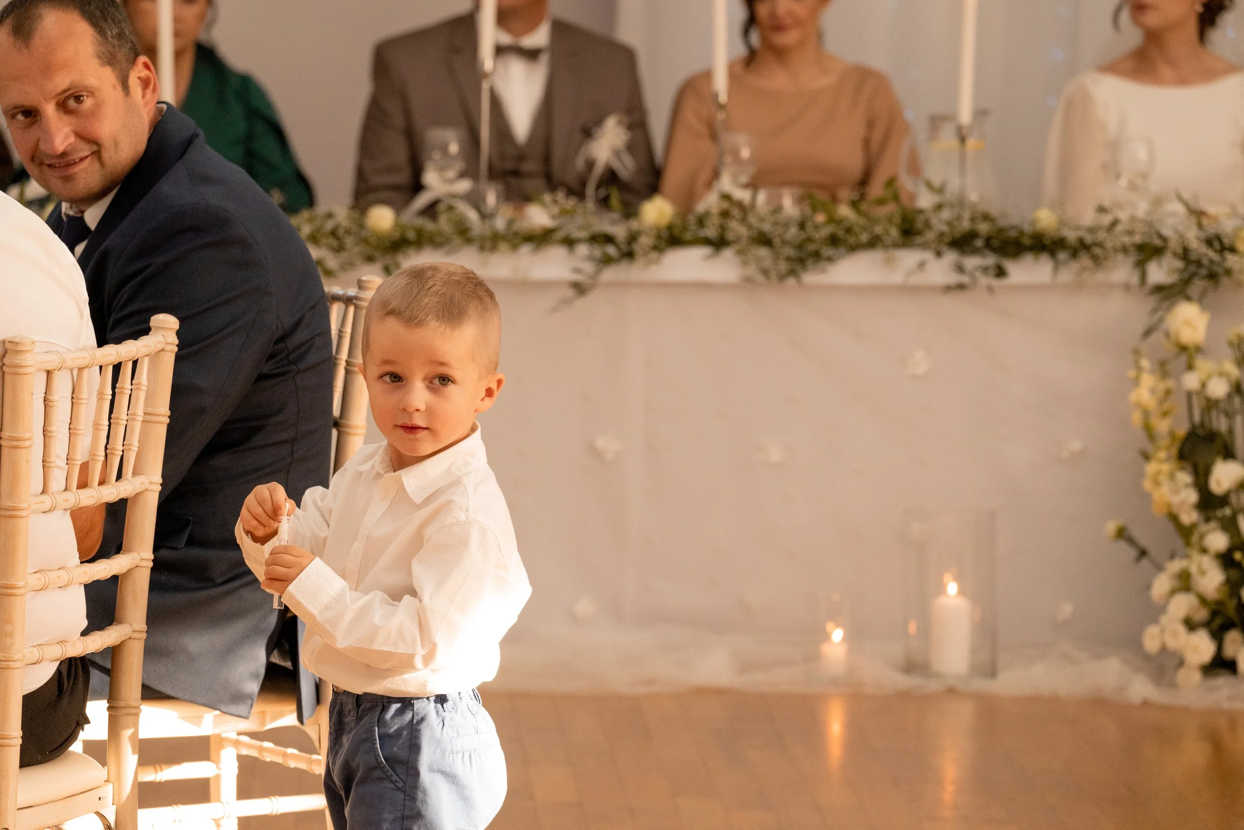 A young boy with short blonde hair wearing a white shirt and jeans, standing at a formal event, looking towards the camera. In the background, guests are seated at a table decorated with candles and flowers. Photographed by Andrea Cabajova, Girl with