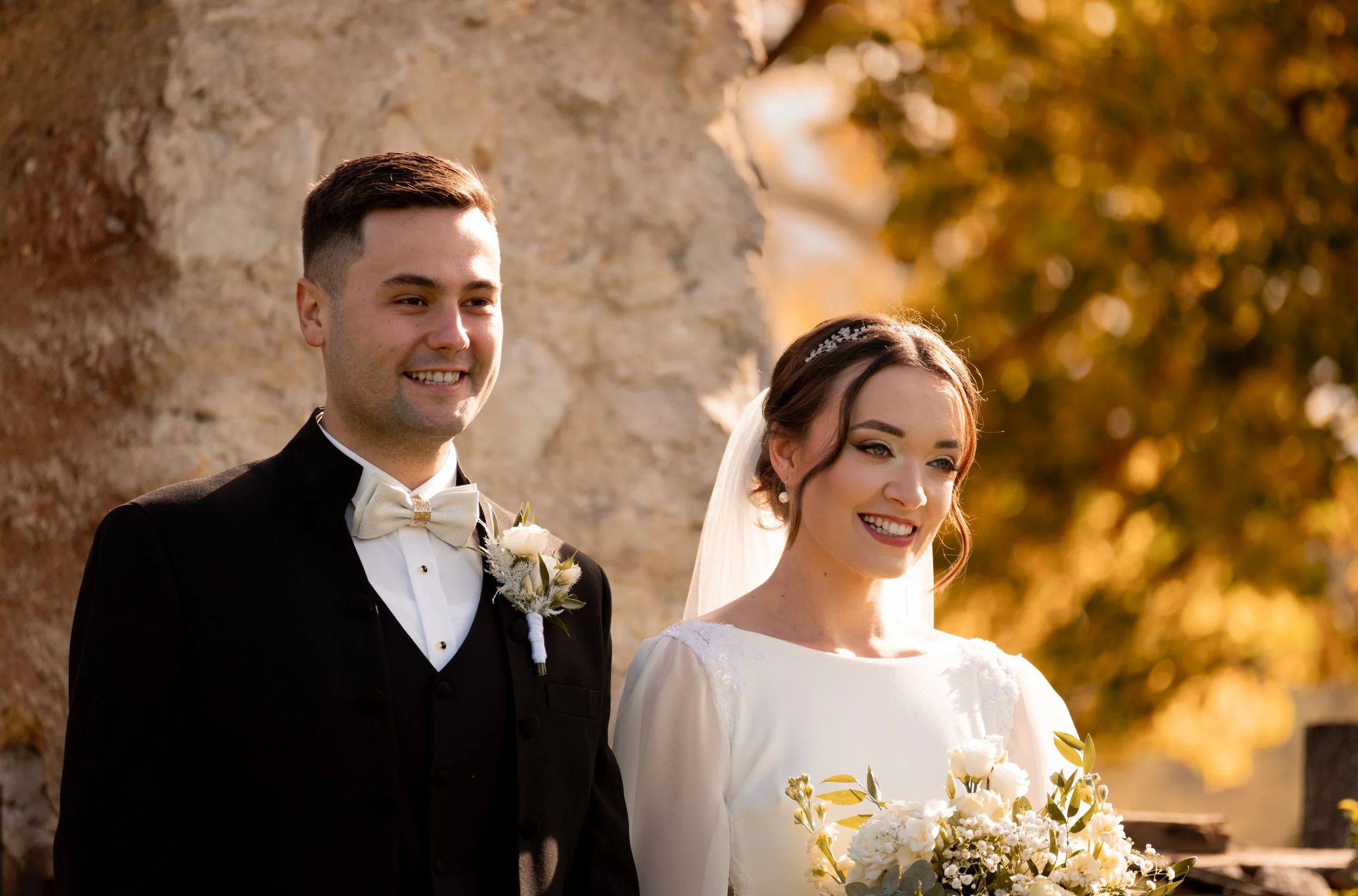 A bride and groom standing outdoors, smiling, during a wedding ceremony with autumn trees in the background. Photographed by Andrea Cabajova, Girl with Bandana.