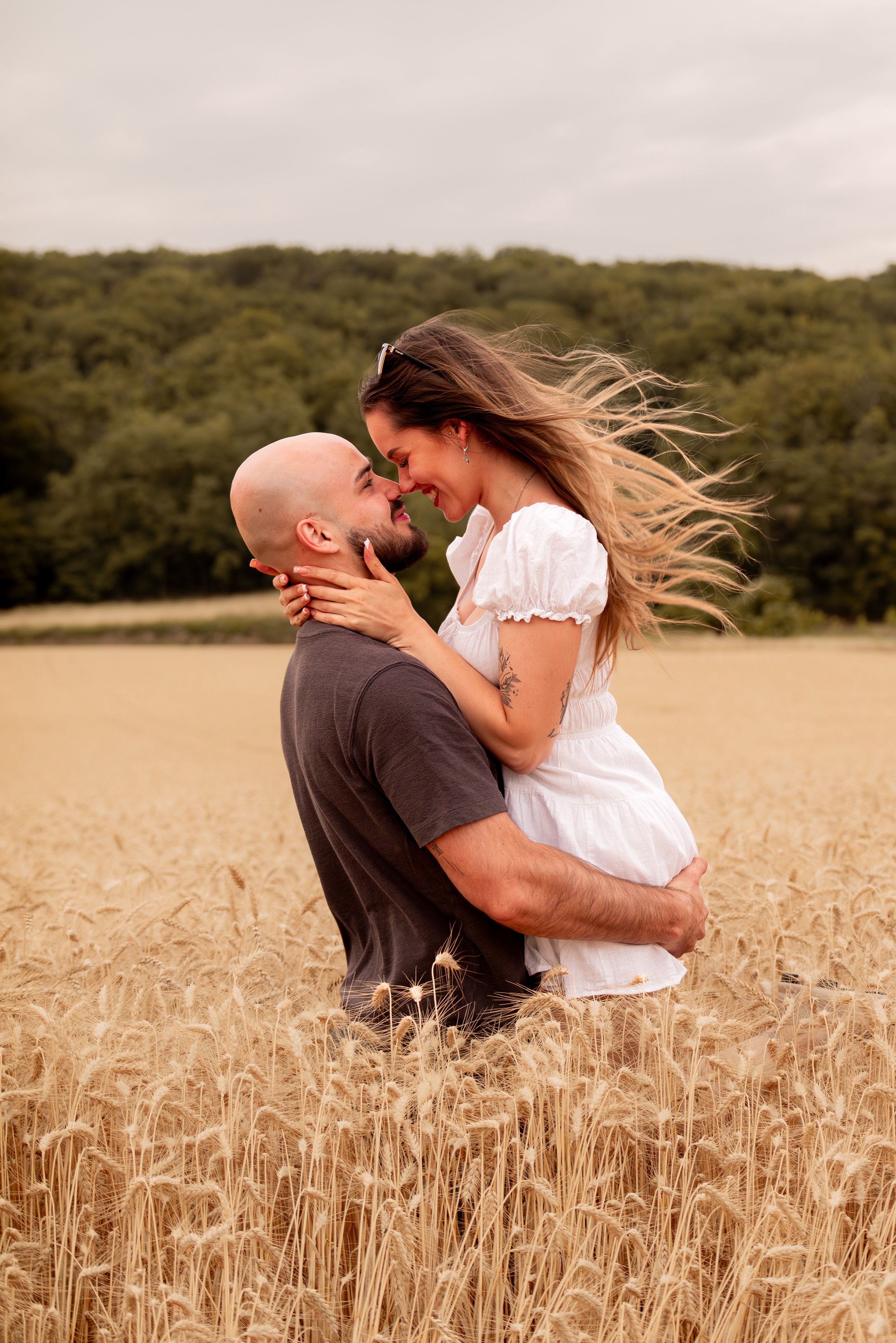 A couple embraces in a wheat field with a forest in the background and a cloudy sky.