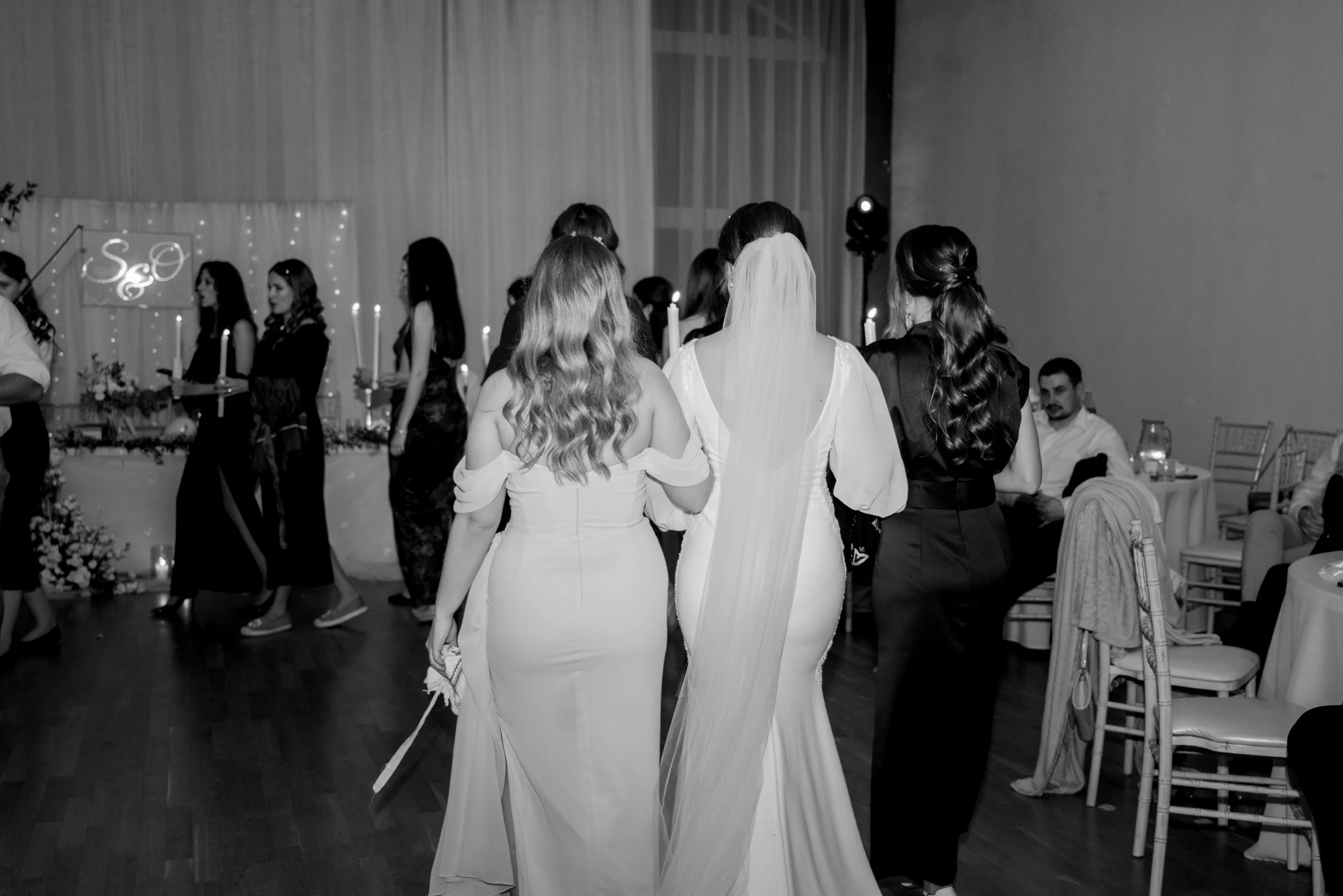 Black and white photo of a wedding reception, with three women in wedding dresses/tuxedos walking together and other guests in the background. Photographed by Andrea Cabajova, Girl with Bandana.