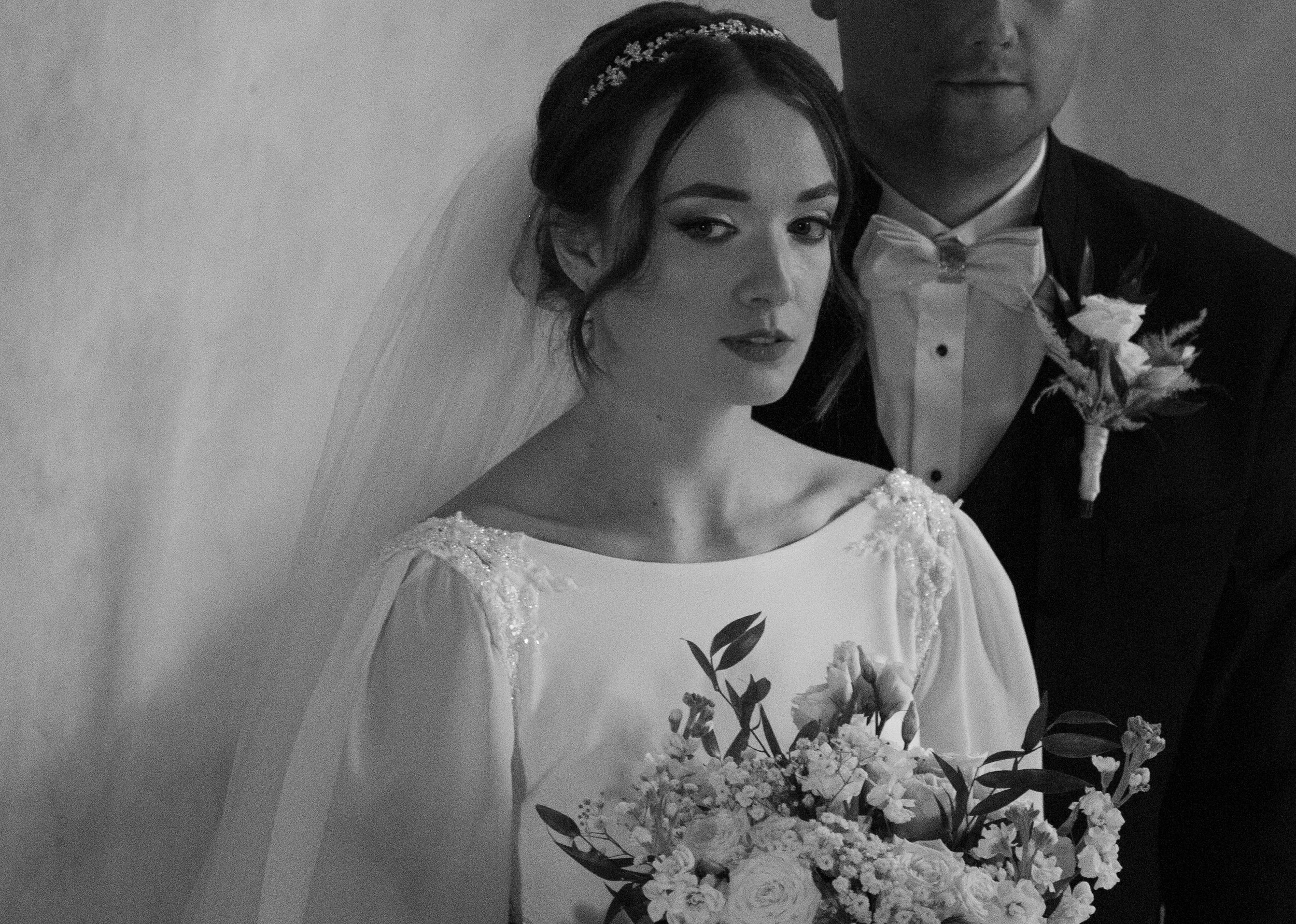 Black and white photo of a bride holding a bouquet of flowers, standing beside a groom in a tuxedo with a boutonniere. Photographed by Andrea Cabajova, Girl with Bandana.