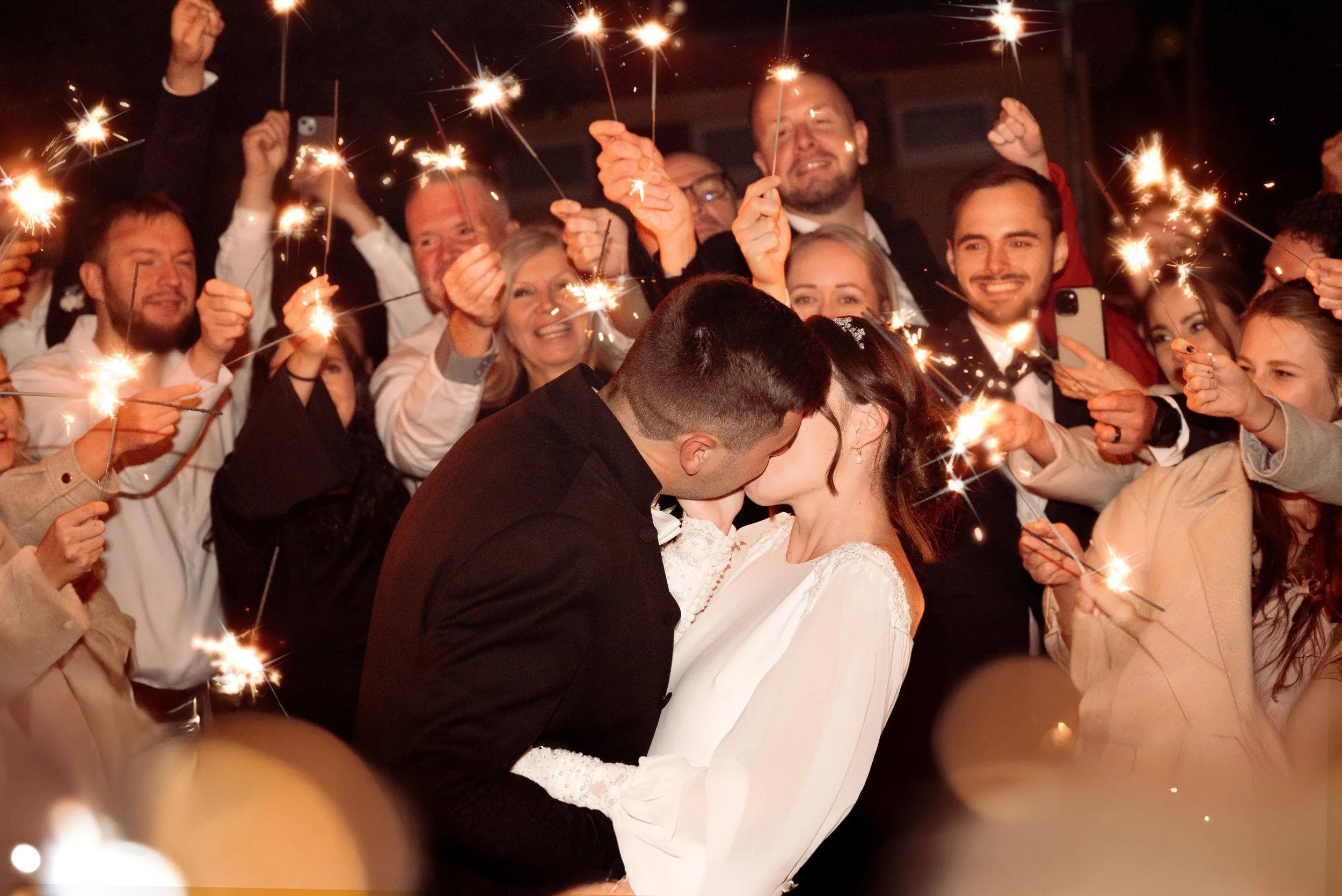 A bride and groom kiss with sparklers held by friends and family surrounding them at night.