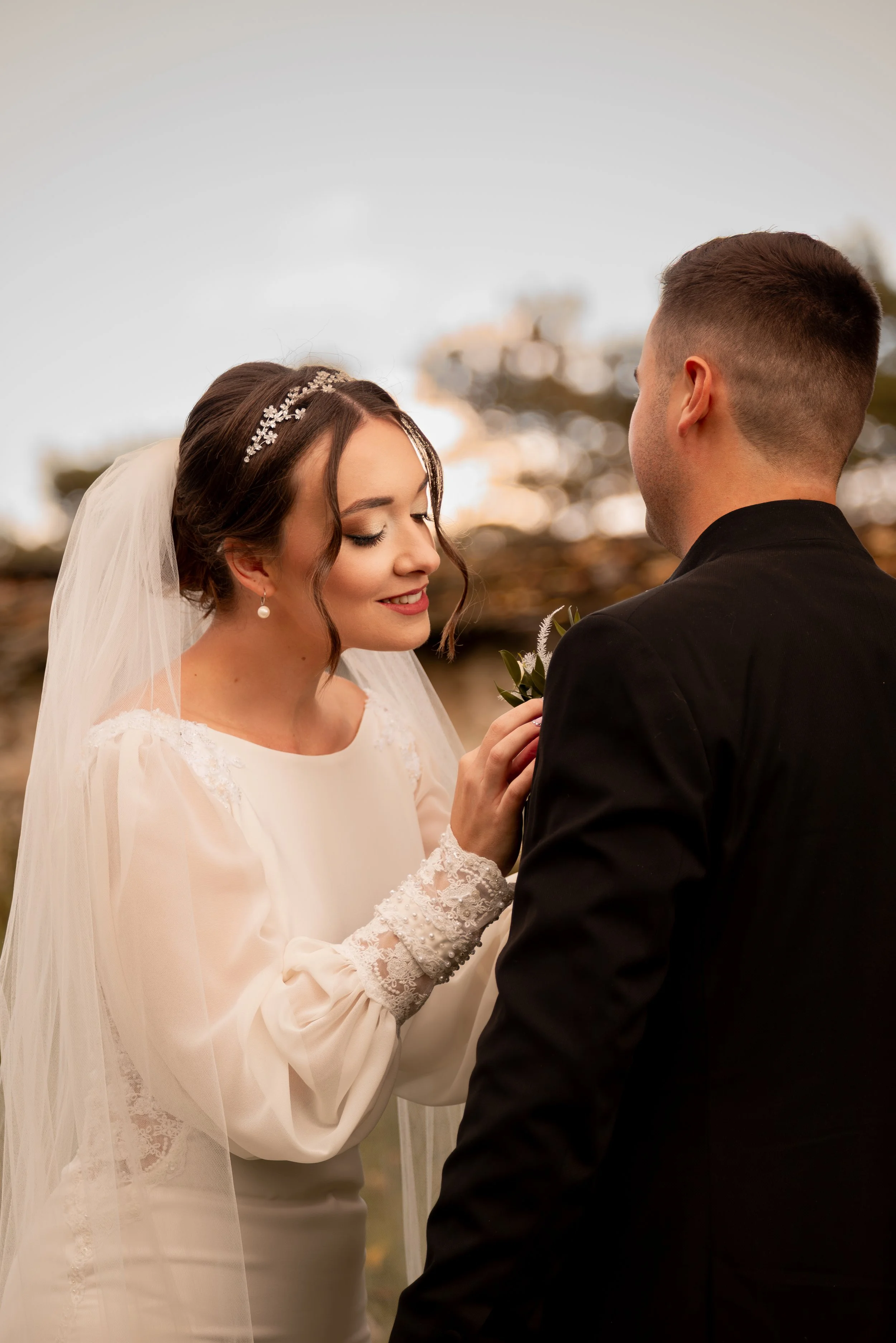 A bride and groom sharing a moment outdoors during sunset. The bride is smiling with eyes closed, wearing a white wedding dress with lace detail on the sleeves and a veil, and holding a small bouquet. Photographed by Andrea Cabajova, Girl with Bandan