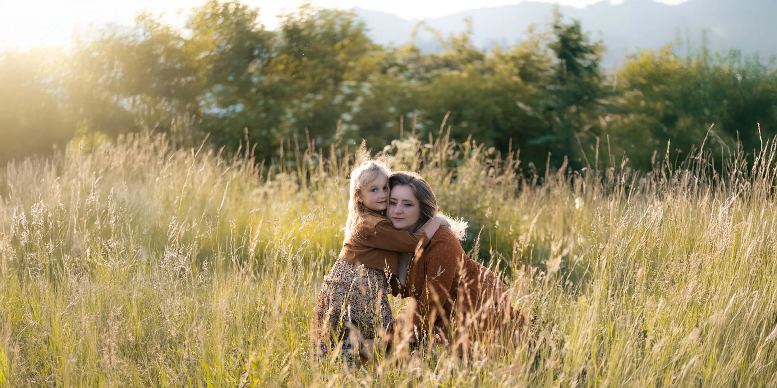 A woman and a young girl embrace in a field of tall grass during sunset, with trees in the background. Photographed by Andrea Cabajova, Girl with Bandana.