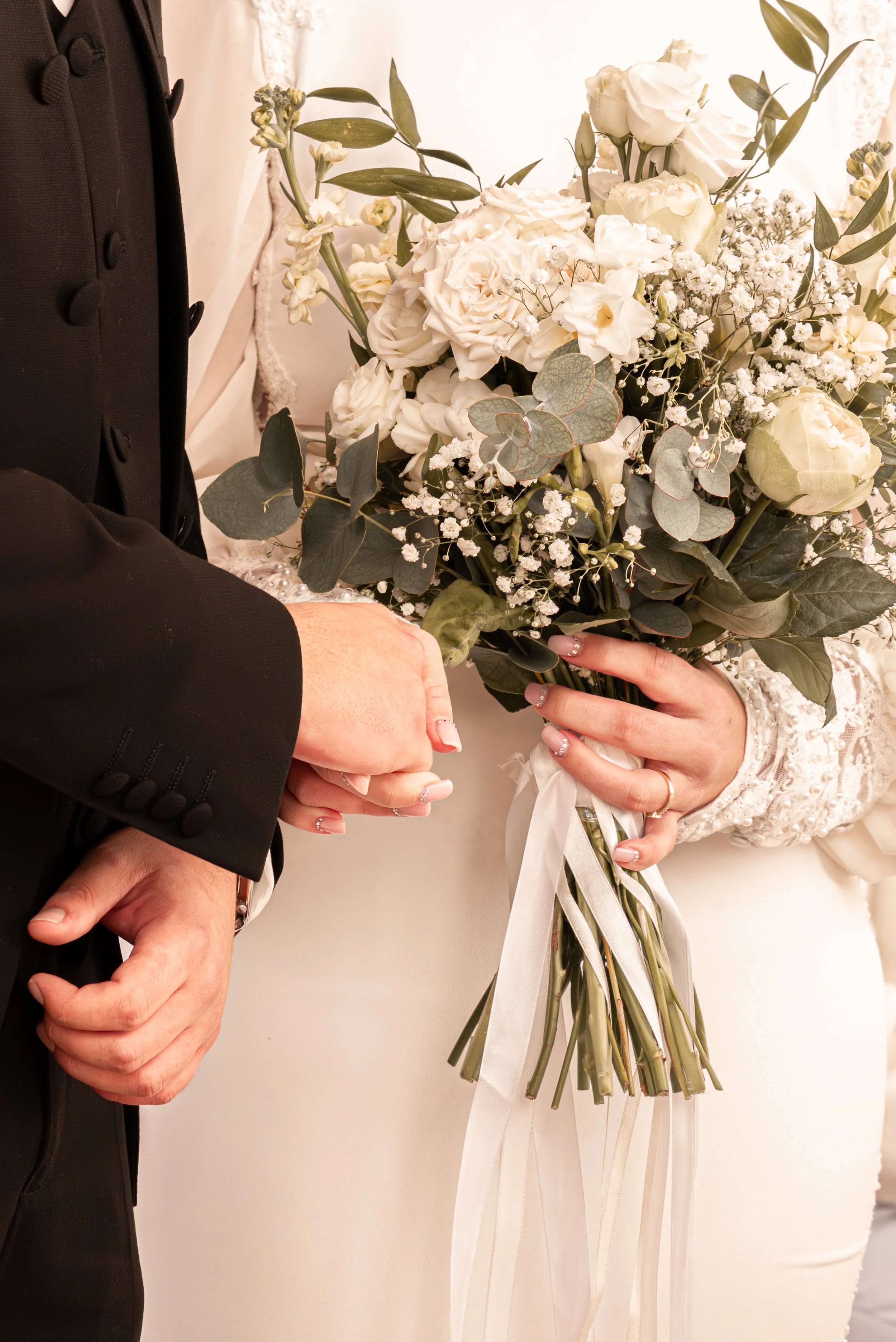 Close-up of a bride and groom holding a bouquet of white flowers and greenery, with the bride's hand featuring a wedding ring. Photographed by Andrea Cabajova, Girl with Bandana.