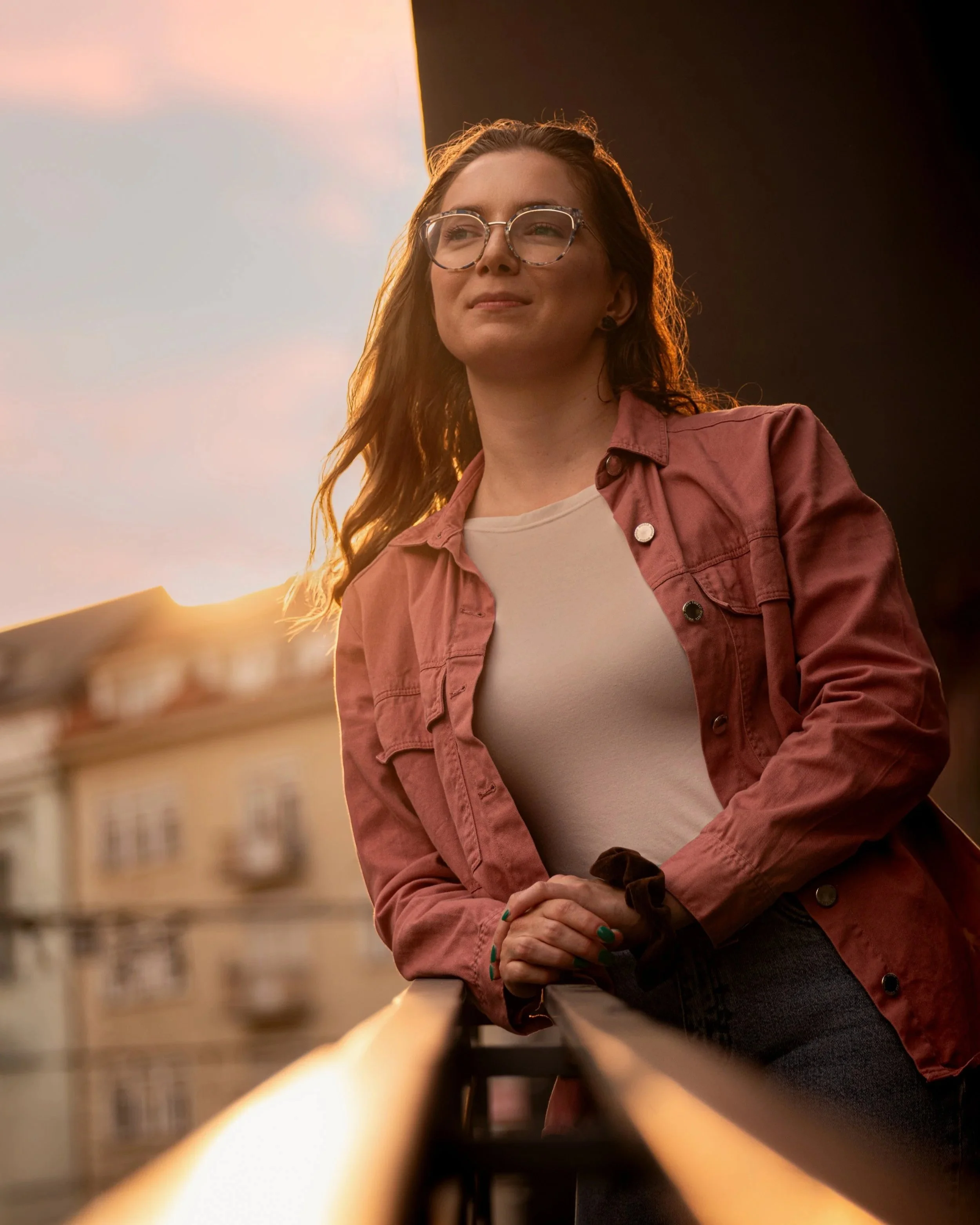 A woman with long red hair and glasses leaning on a balcony railing at sunset, wearing a pink jacket and a white shirt. Photographed by Andrea Cabajova, Girl with Bandana.