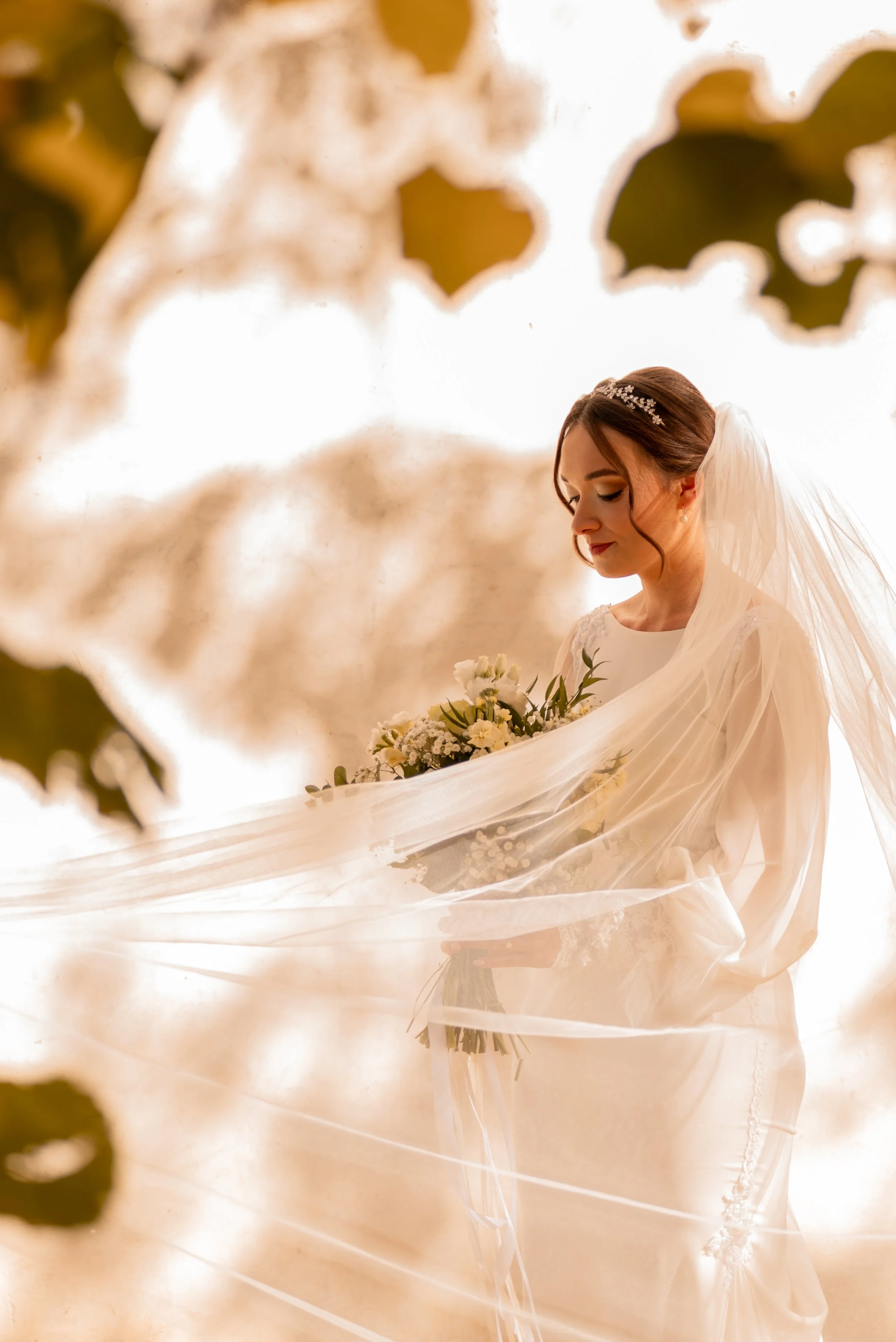 A bride holding a bouquet, dressed in a white wedding gown with a sheer veil, stands with eyes closed and a gentle smile, surrounded by soft, warm lighting and blurred green leaves in the foreground. Photographed by Andrea Cabajova, Girl with Bandana