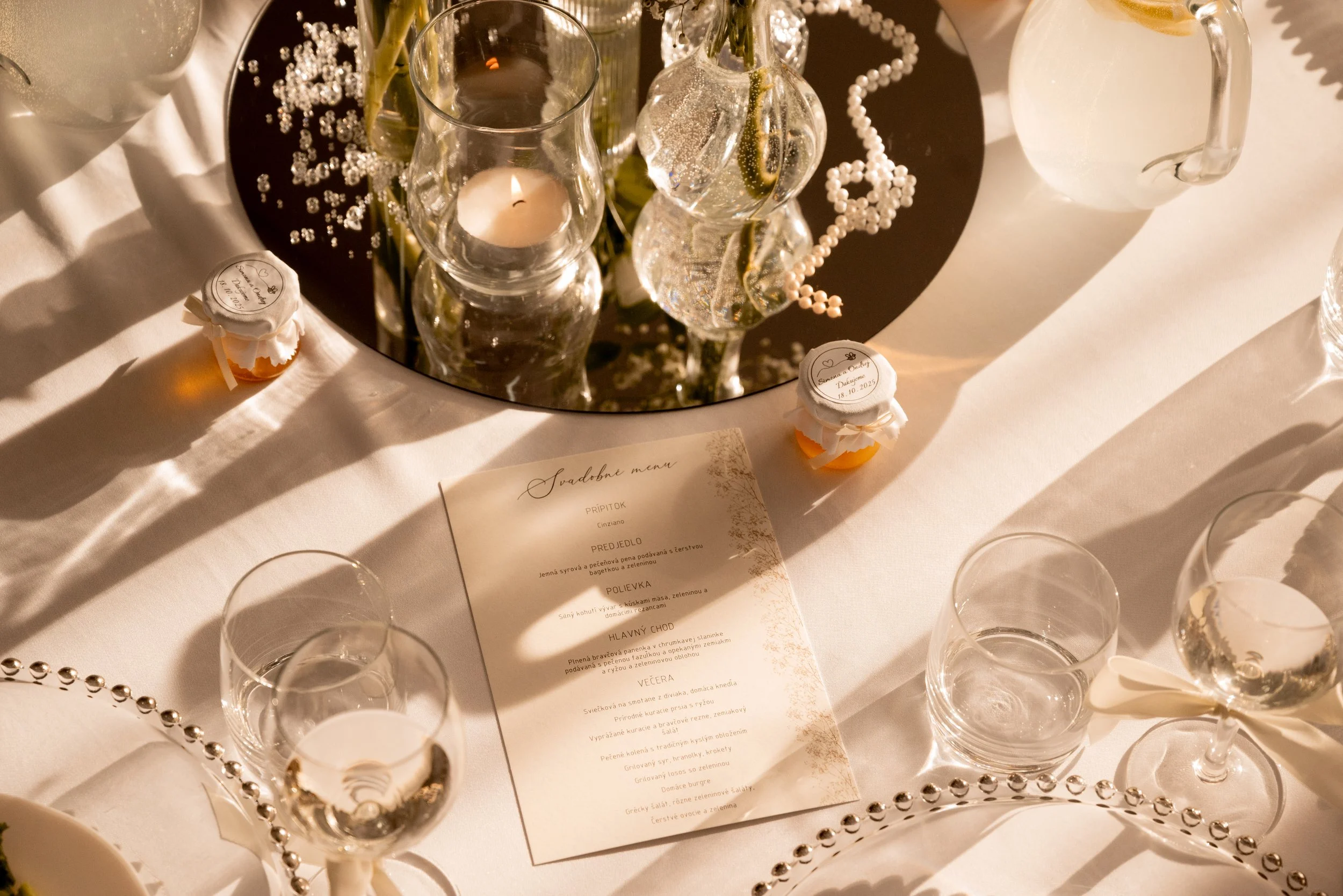 Elegant table setting with a menu, glasses, a mirror centerpiece with candles, small wrapped favors, and decorative pearls, suggesting a formal event or celebration.