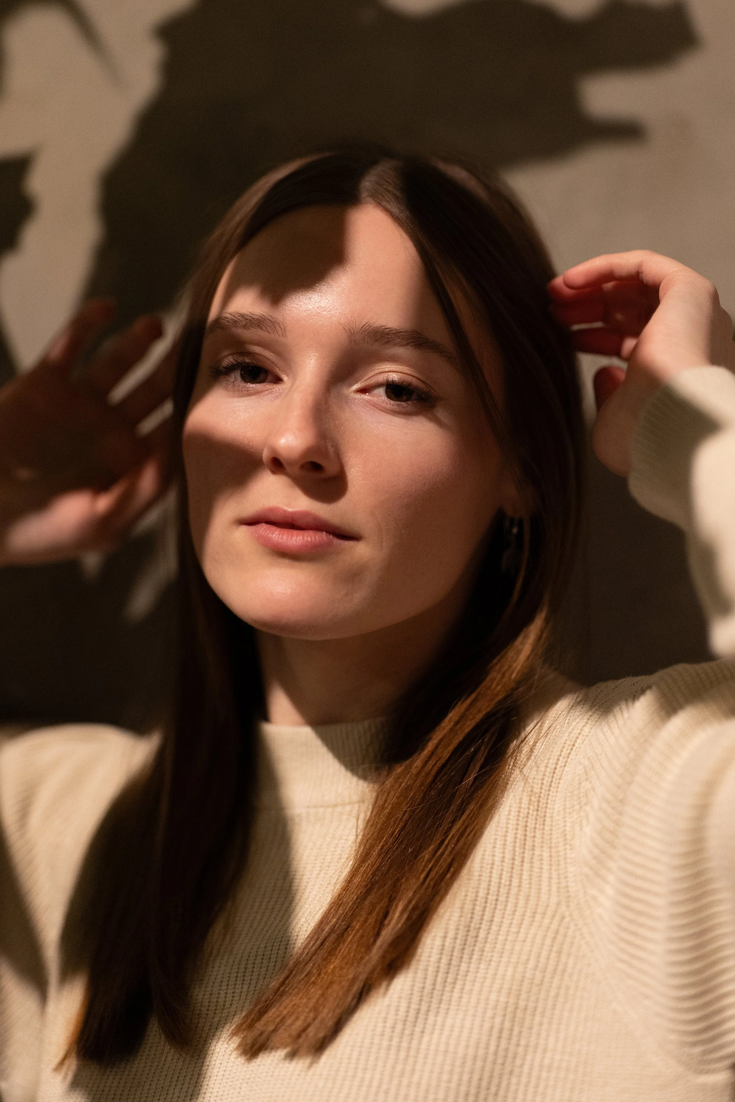 A young woman with brown hair wearing a beige sweater, posing with her hands near her head, casting shadows on her face against a dark background. Photographed by Andrea Cabajova, Girl with Bandana.