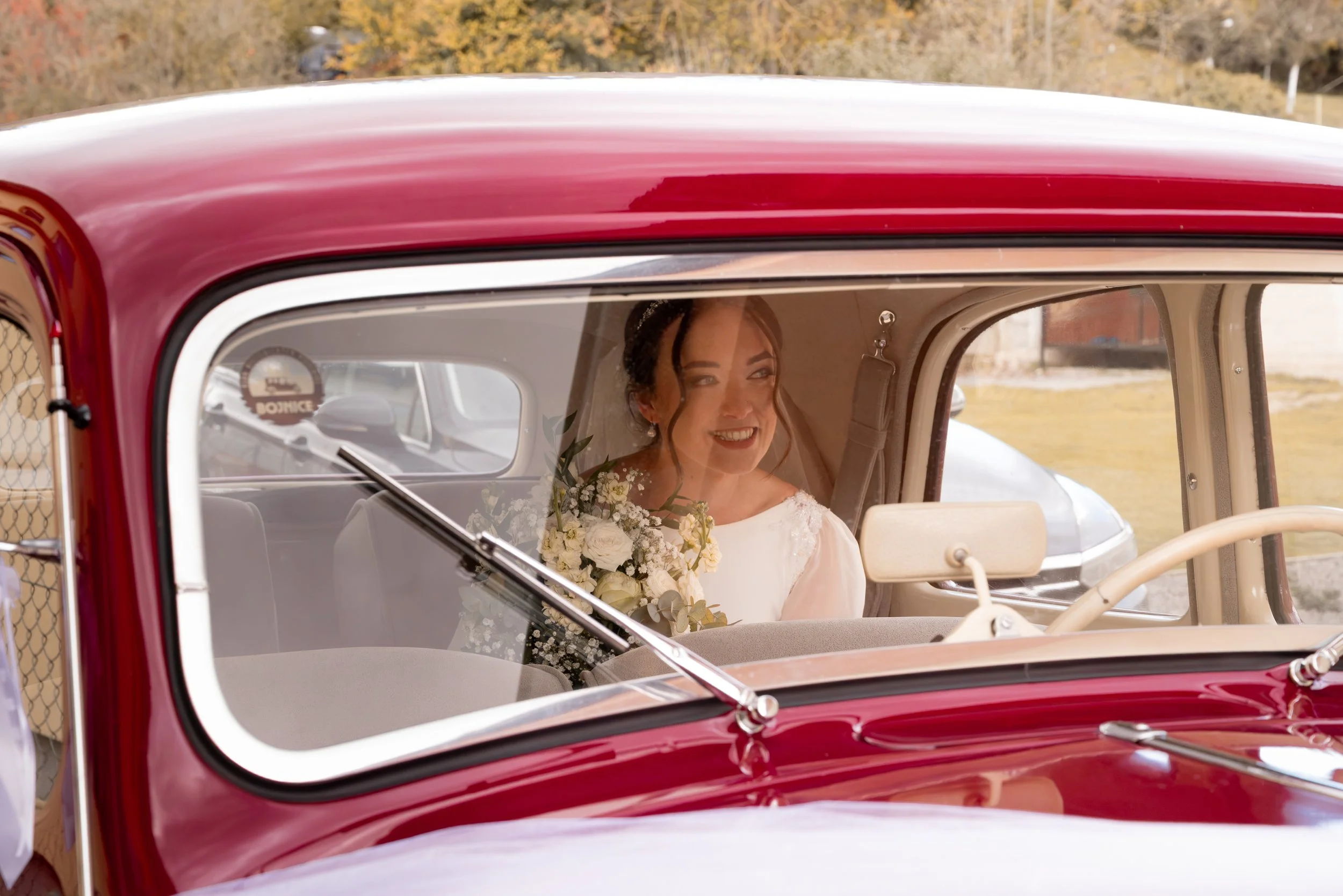 A bride sitting inside a vintage red car, holding a bouquet of white flowers and smiling. Photographed by Andrea Cabajova, Girl with Bandana.