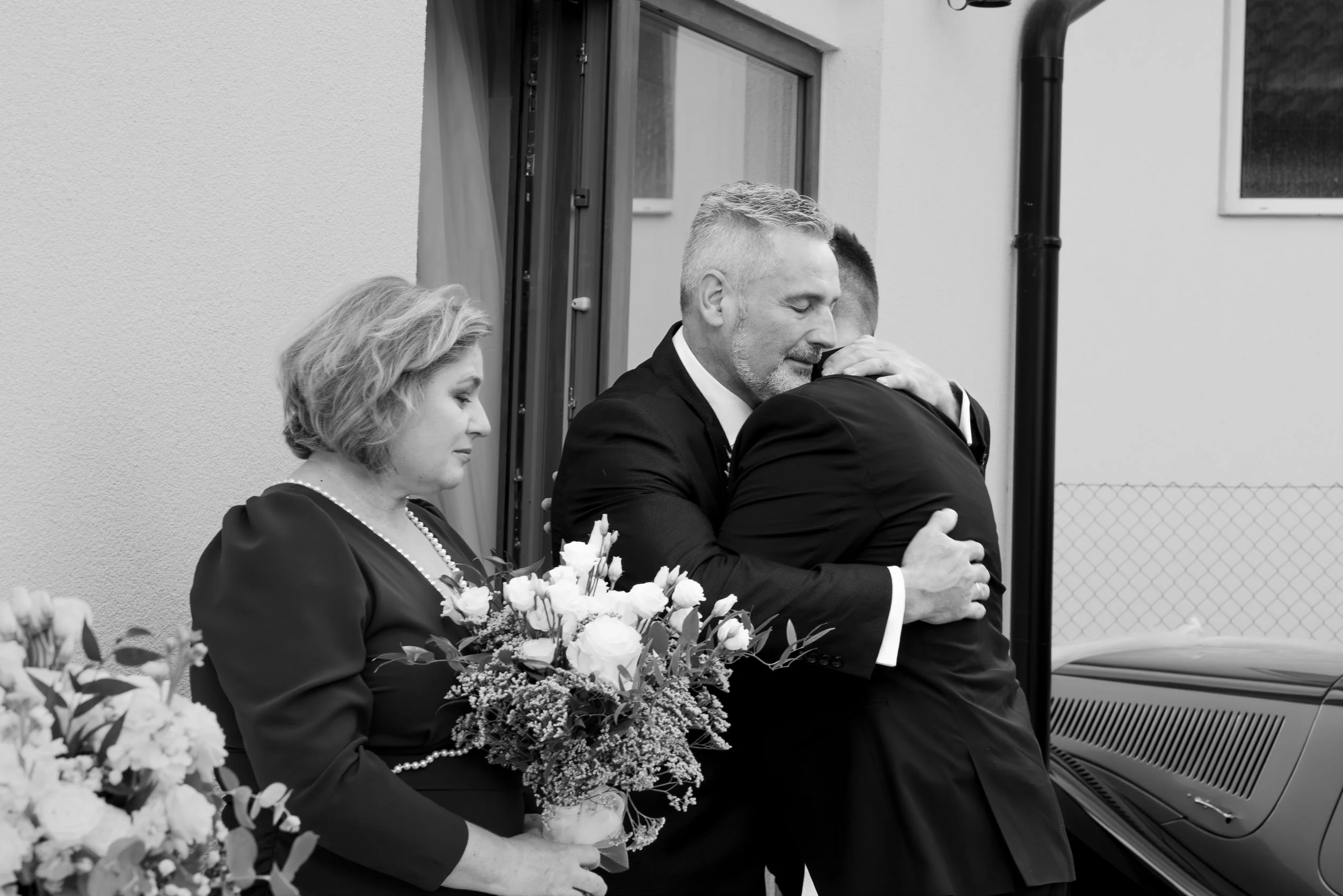 A black and white photo of a family embracing at a celebration, with a woman holding a bouquet of flowers and a man hugging someone, standing outside a building with a window and fence in the background. Photographed by Andrea Cabajova, Girl with Ban