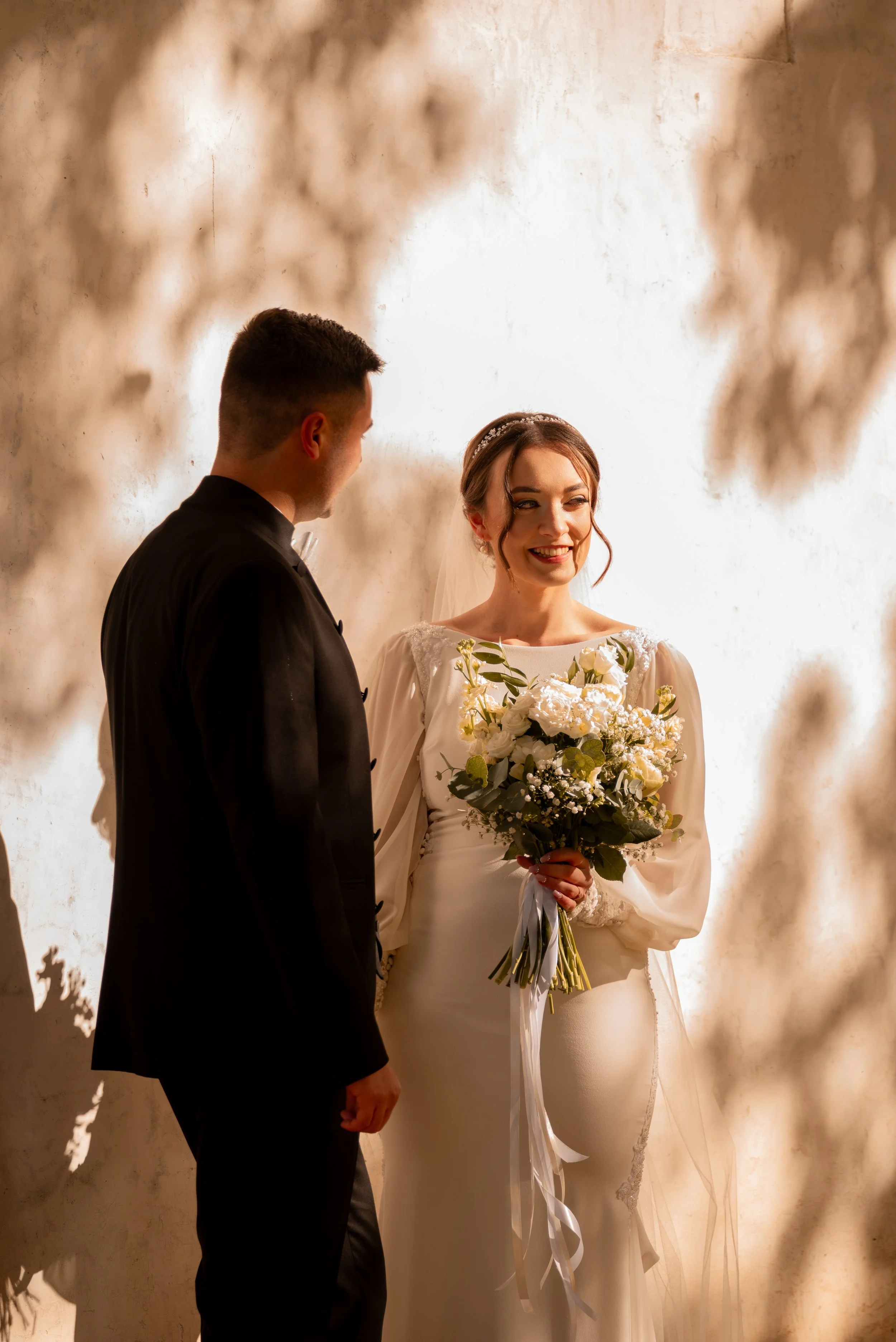 A bride and groom on their wedding day standing by a textured wall, with the bride holding a bouquet of white flowers and greenery, smiling at each other. Photographed by Andrea Cabajova, Girl with Bandana.