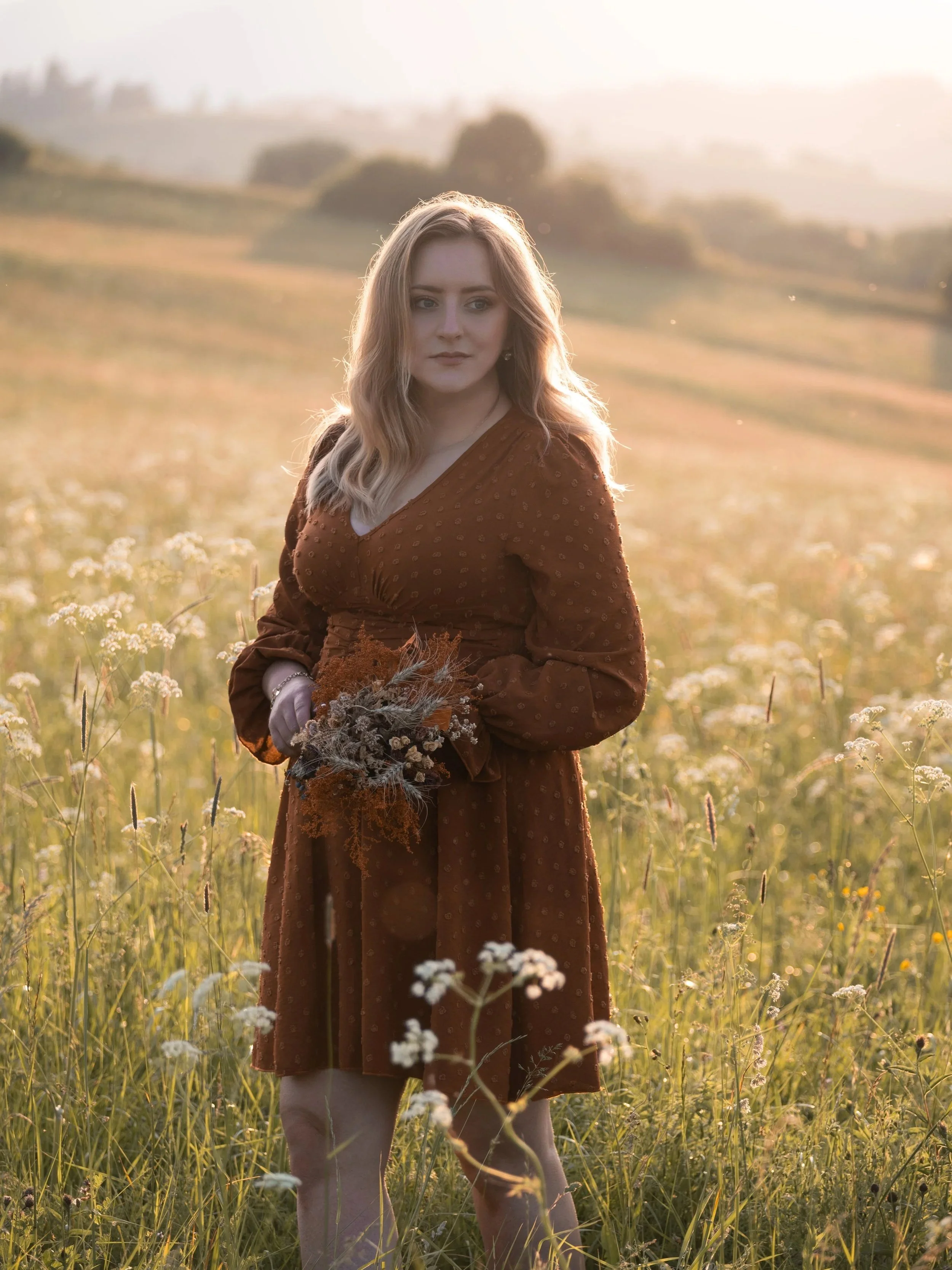 A woman in a brown dress holding a bouquet of dried flowers standing in a field of tall grass and flowers during sunset. Photographed by Andrea Cabajova, Girl with Bandana.