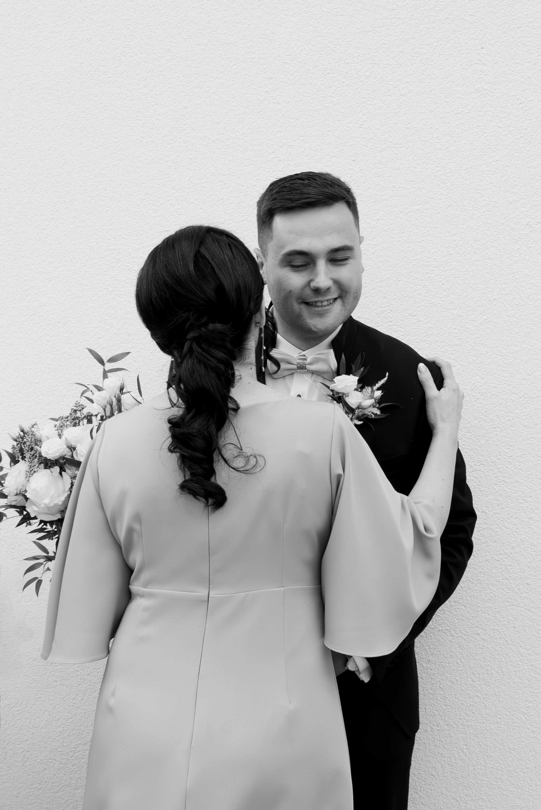 A couple at a wedding, with the woman holding a bouquet and the man dressed in a tuxedo with a boutonniere, sharing a close moment against a plain wall. Photographed by Andrea Cabajova, Girl with Bandana.