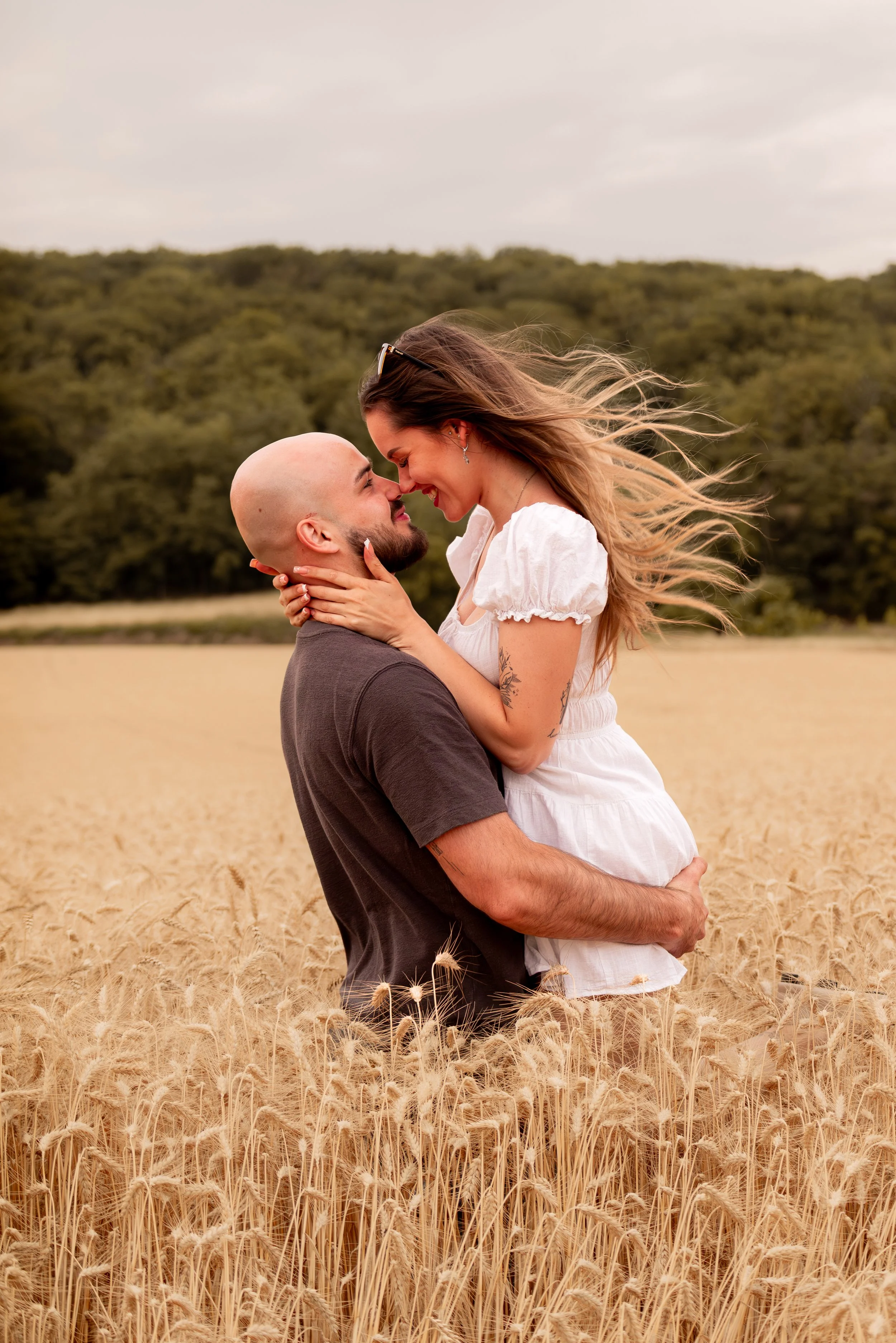 A couple embracing in a wheat field with a backdrop of trees and cloudy sky, their faces close together, smiling. Photographed by Andrea Cabajova, Girl with Bandana.