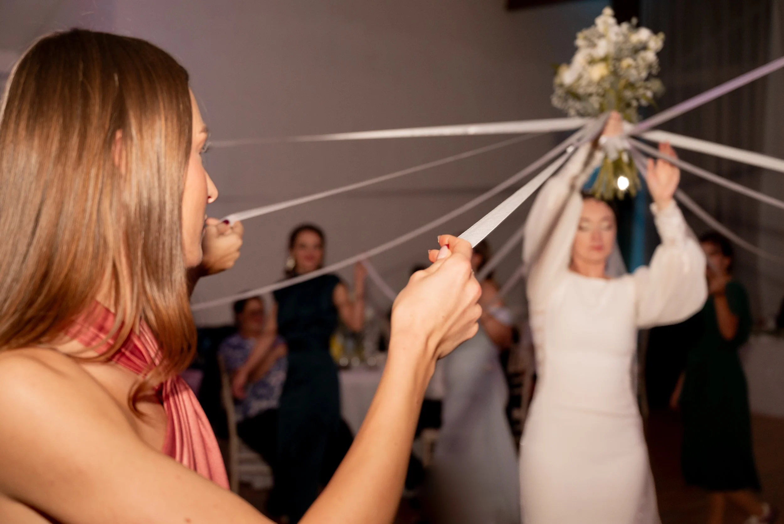 Women participating in a bouquet ceremony where ribbons are attached to a bouquet held overhead, with guests observing.