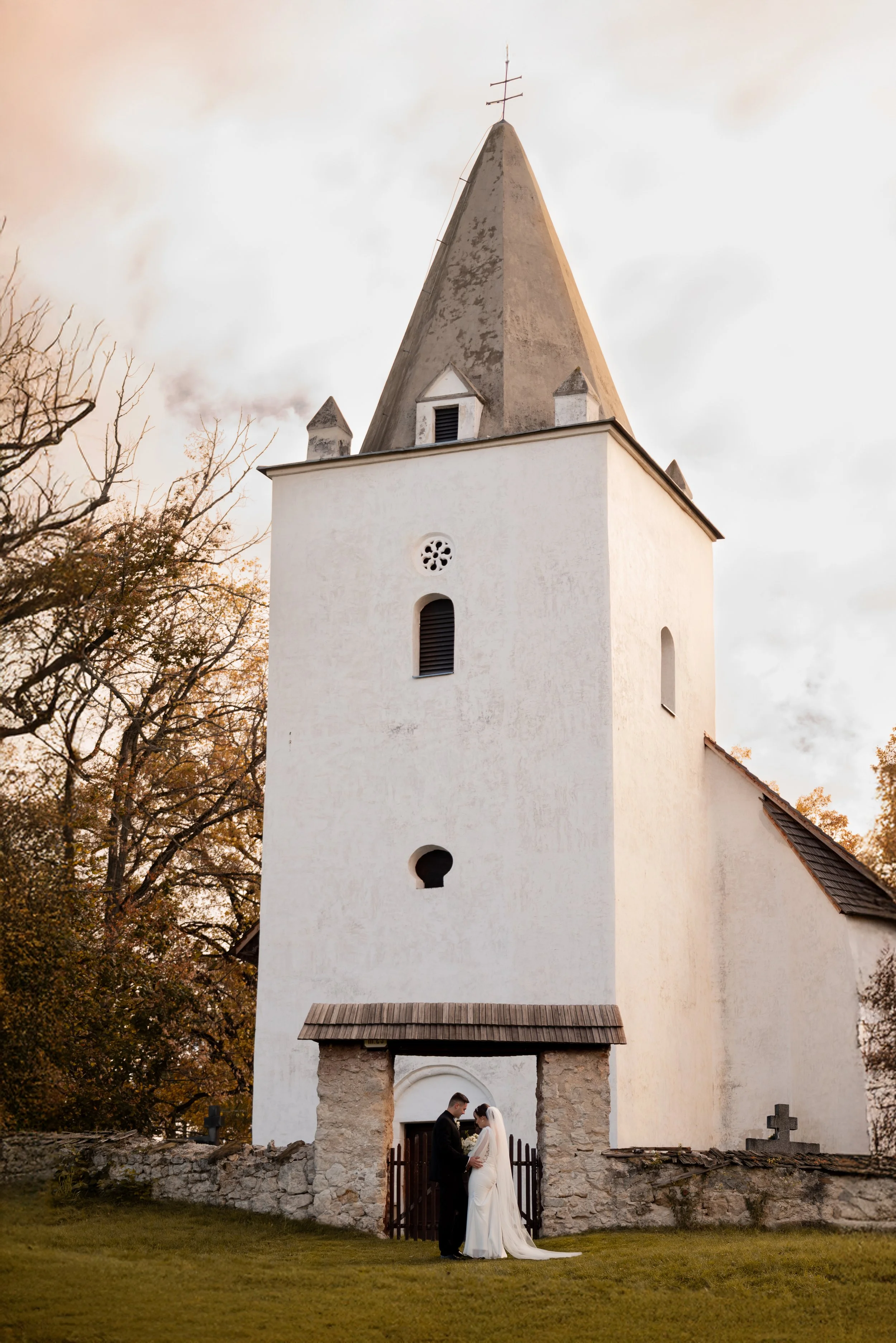 A bride and groom standing together in front of a small church with a tall steeple, holding hands, during sunset. Photographed by Andrea Cabajova, Girl with Bandana.