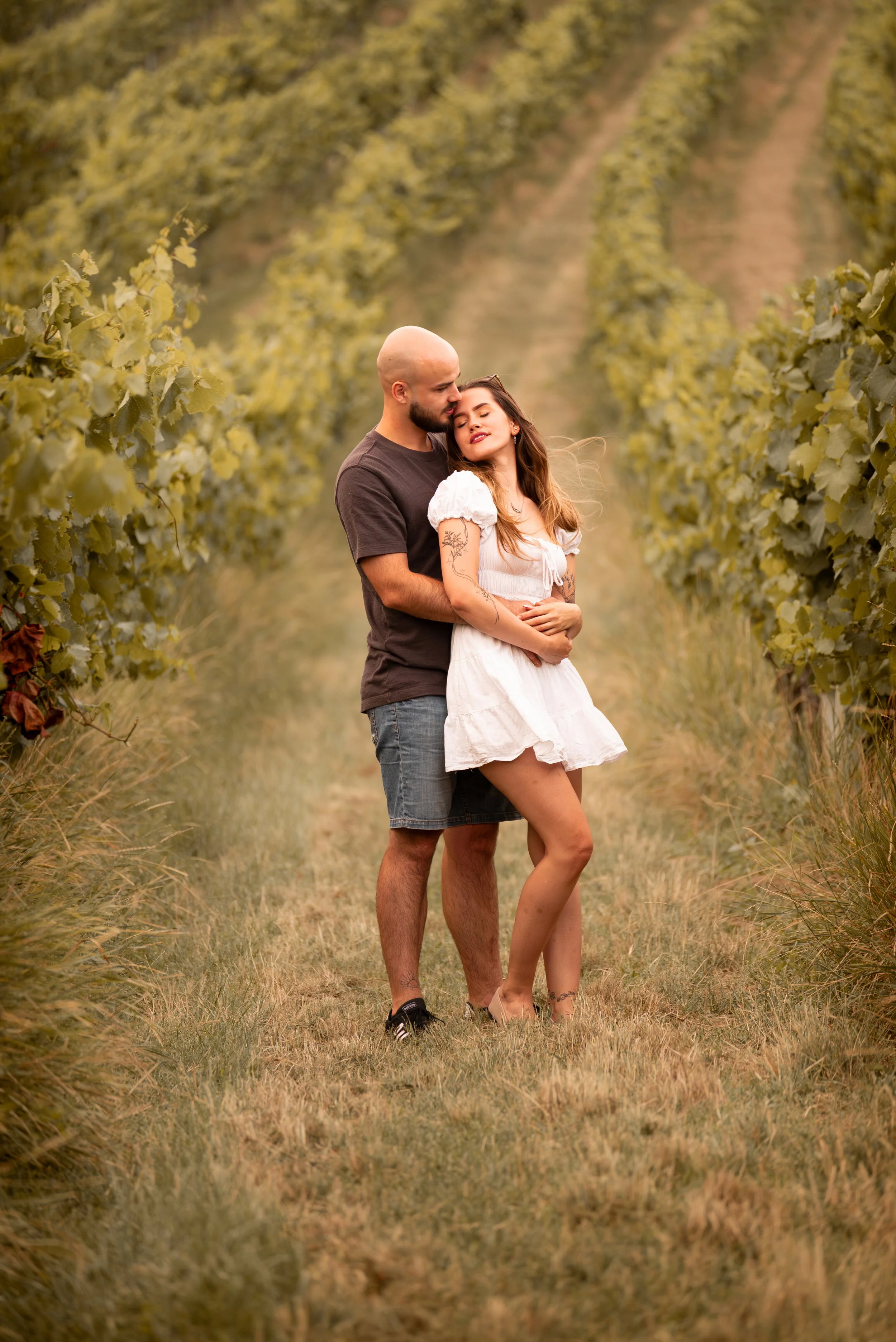 A couple stands close together in a vineyard, with the man behind the woman, embracing her with eyes closed and the woman leaning into him with a serene expression.