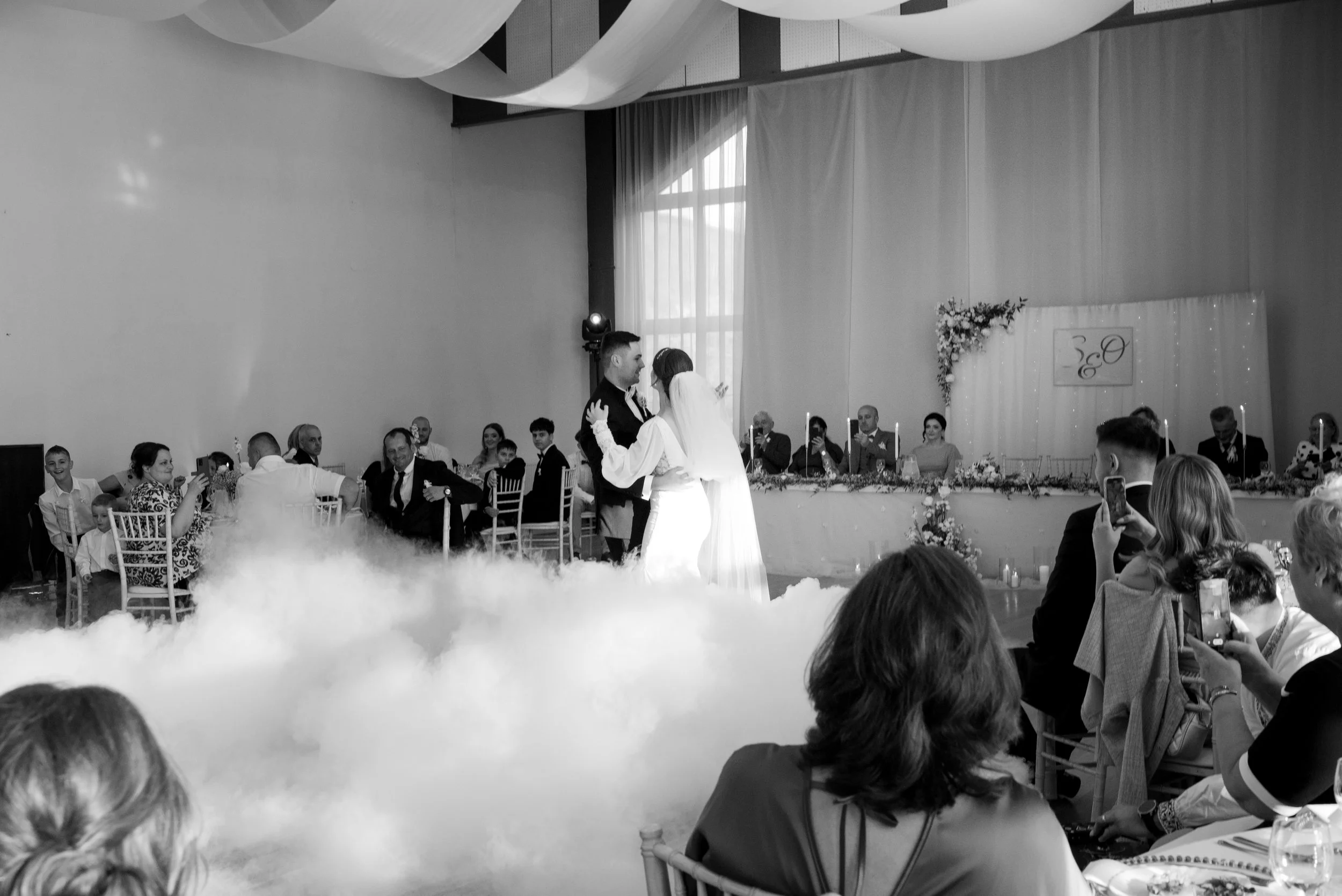 A wedding ceremony with a bride and groom dancing, surrounded by guests, with a decorated backdrop and a long table with seated officials or family members in the background. The scene is filled with people capturing the moment on their phones, and t