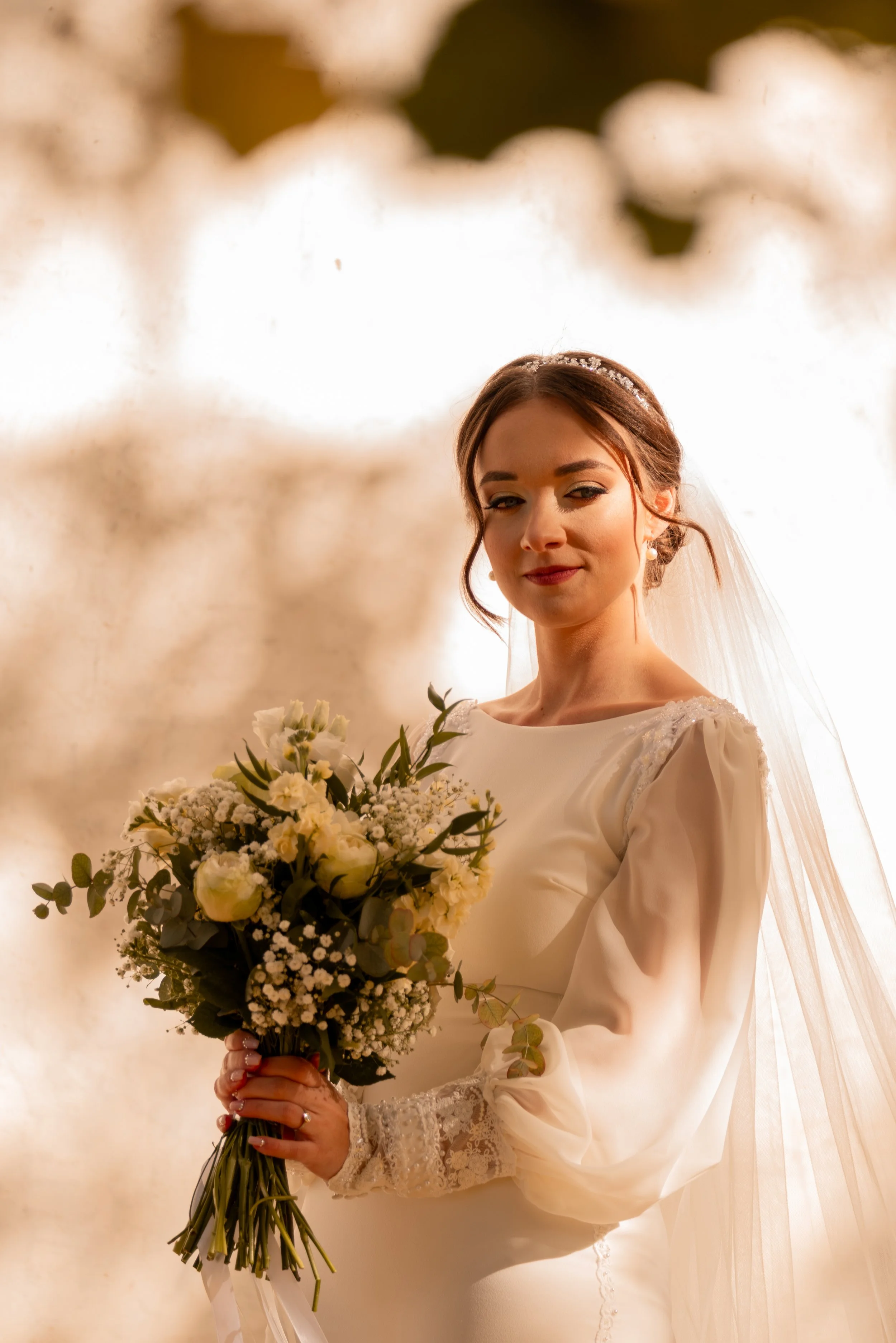 A bride stands in soft lighting, holding a bouquet of white flowers and greenery, with a gentle smile, in a white wedding dress with lace details on the sleeves and a veil. Photographed by Andrea Cabajova, Girl with Bandana.