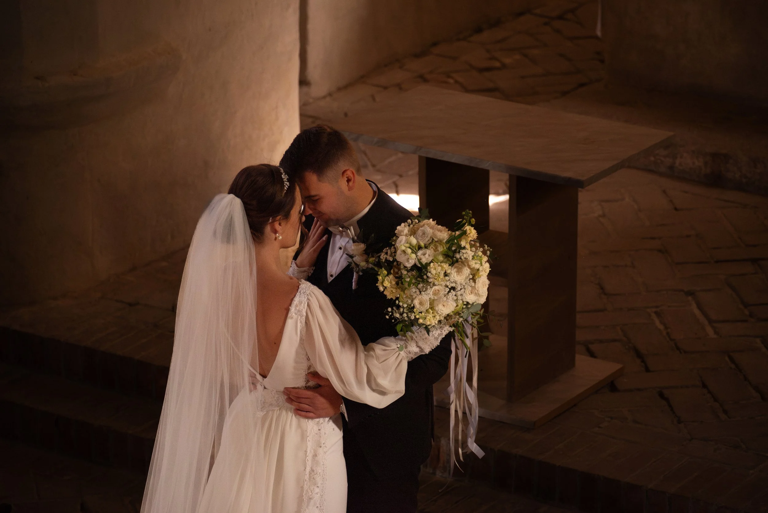 A bride and groom sharing a kiss during their wedding ceremony, with the groom holding a large bouquet of white flowers, in a dimly lit indoor setting. Photographed by Andrea Cabajova, Girl with Bandana.