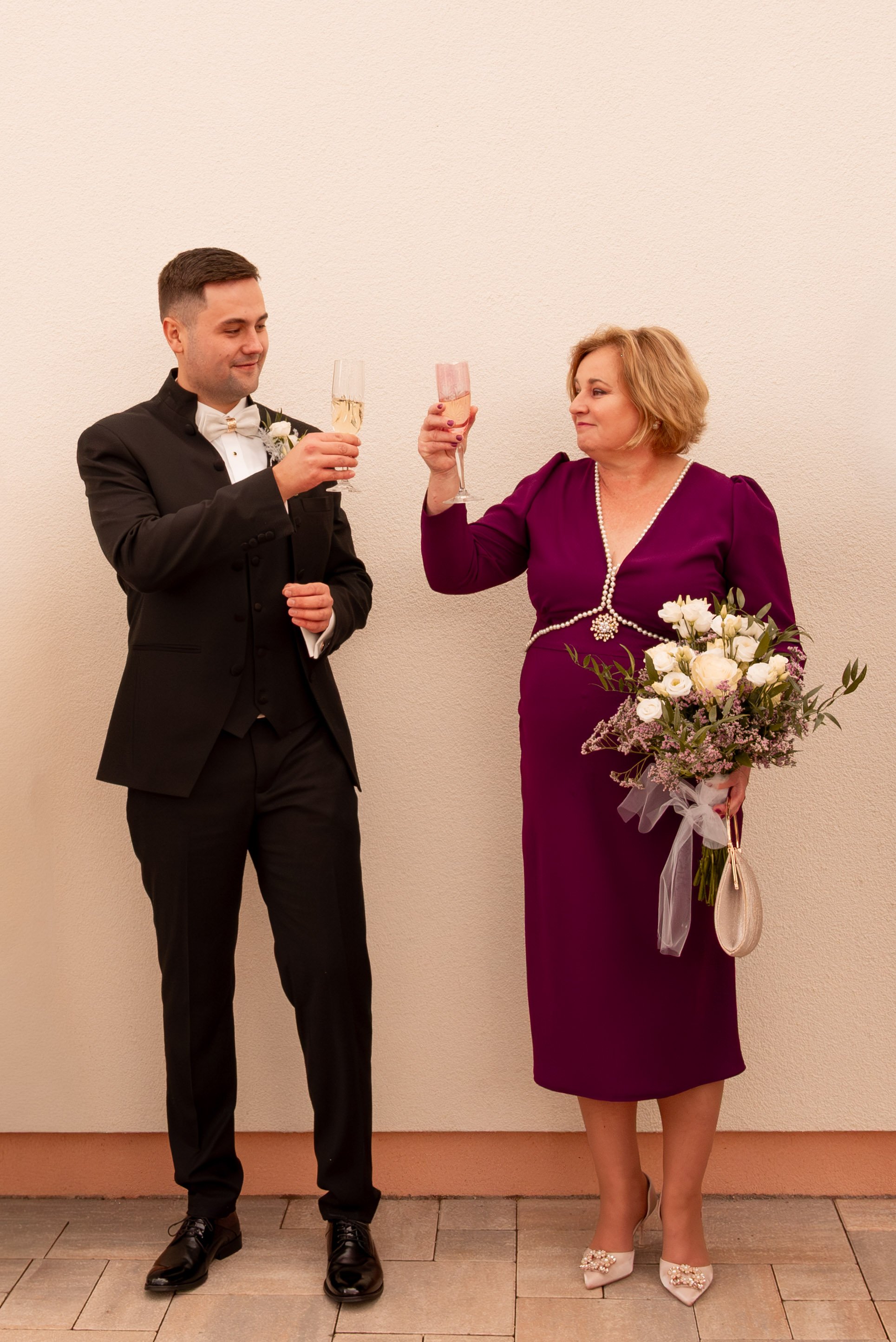 A man in a black suit and woman in a purple dress clink glasses during a celebration, with the woman holding a bouquet of white flowers. Photographed by Andrea Cabajova, Girl with Bandana.