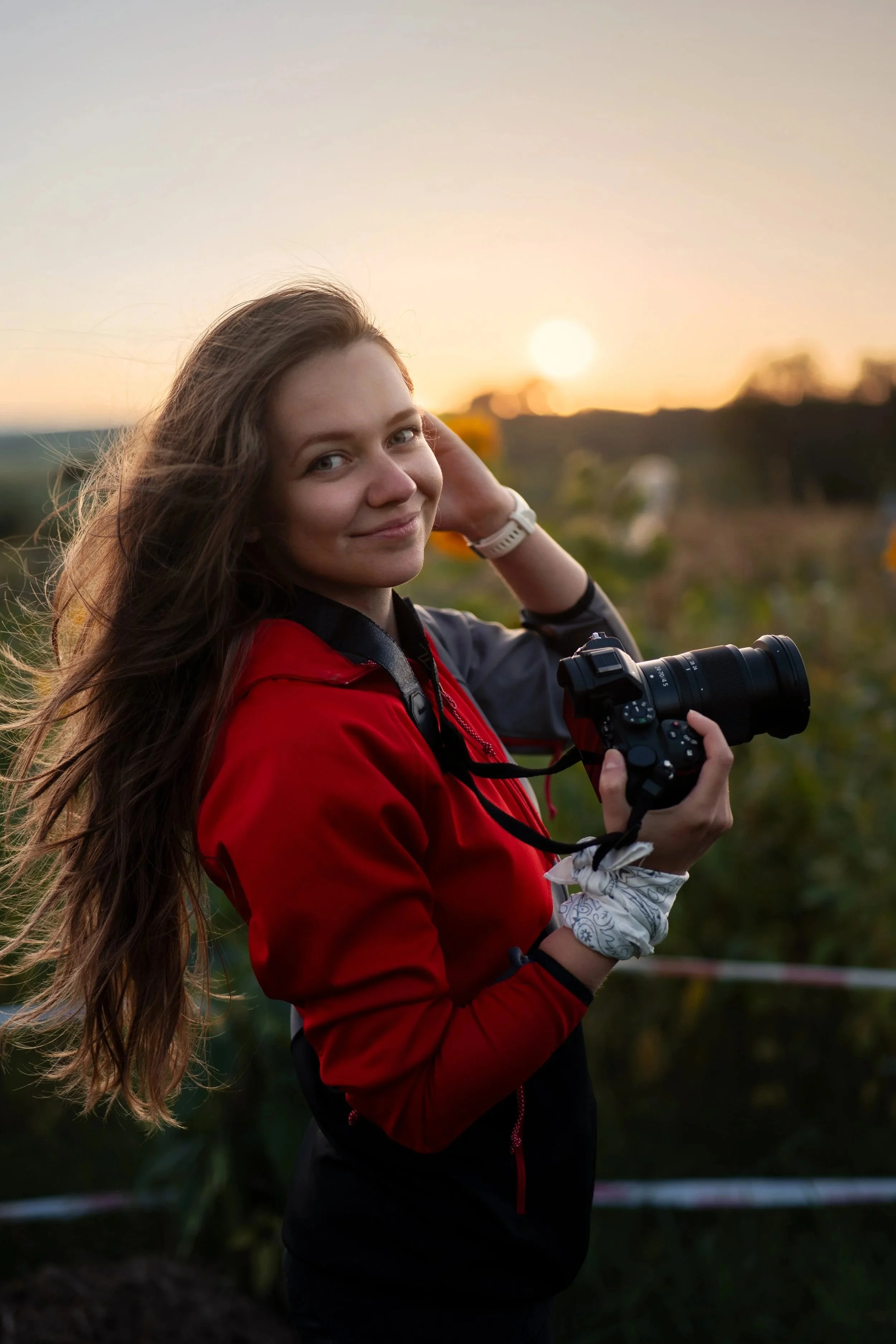 Young woman with long hair smiling while holding a camera outdoors at sunset, wearing a red jacket and a white wristwatch.