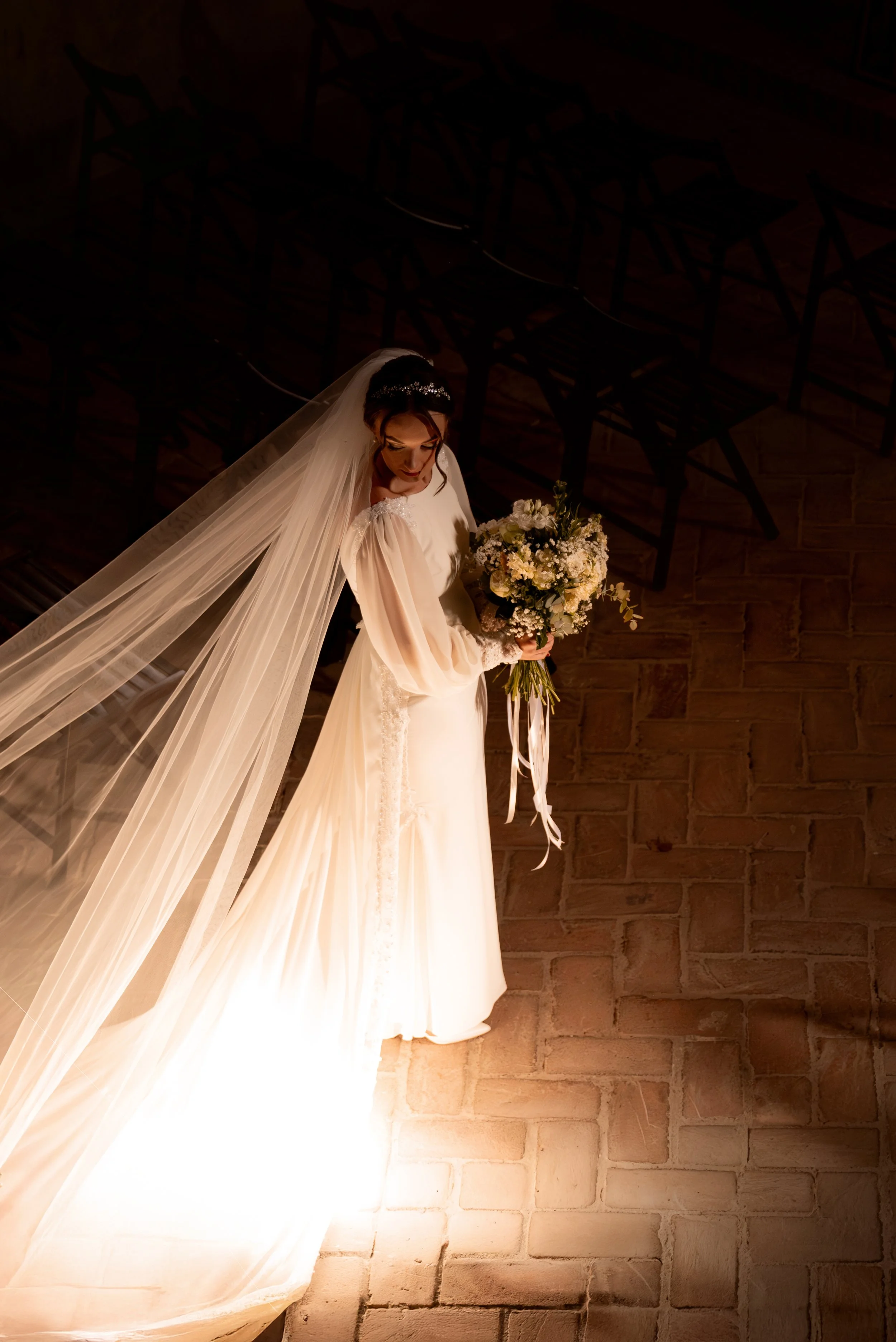 A bride in a white wedding gown and veil holding a bouquet of flowers, standing alone on a lit stage with empty chairs in the background. Photographed by Andrea Cabajova, Girl with Bandana.