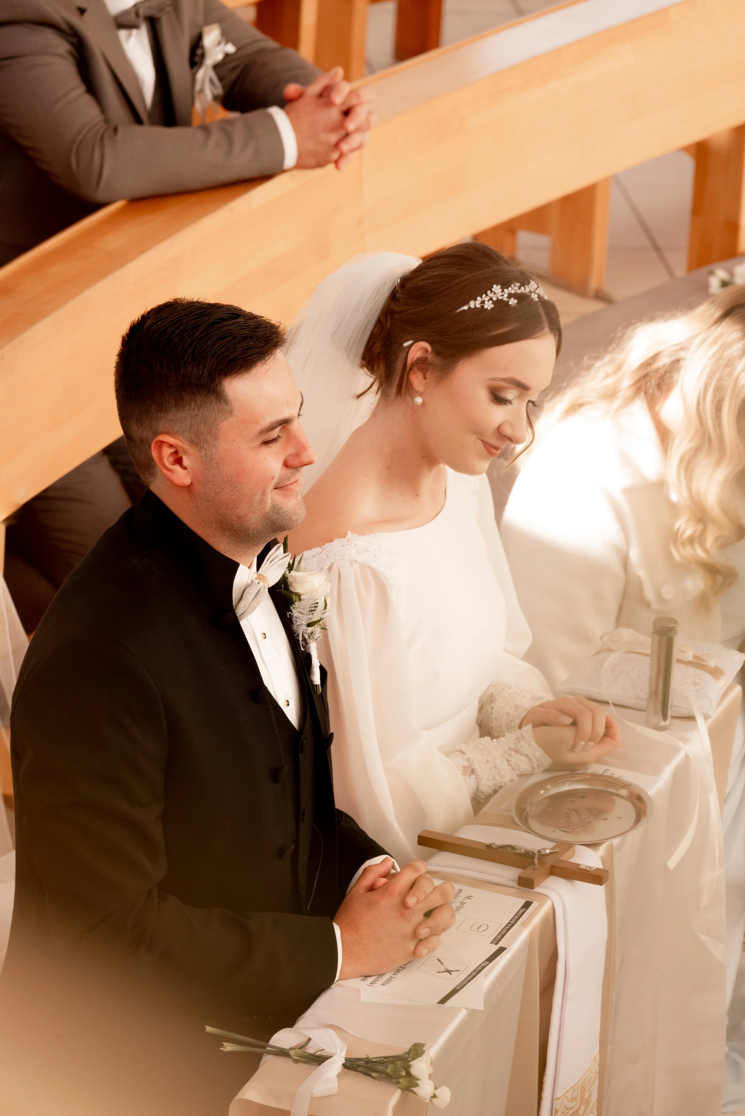A bride and groom kneeling at the altar during a wedding ceremony, with the bride dressed in a white gown and veil, and the groom in a black tuxedo, inside a church with wooden pews and a congregation. Photographed by Andrea Cabajova, Girl with Banda