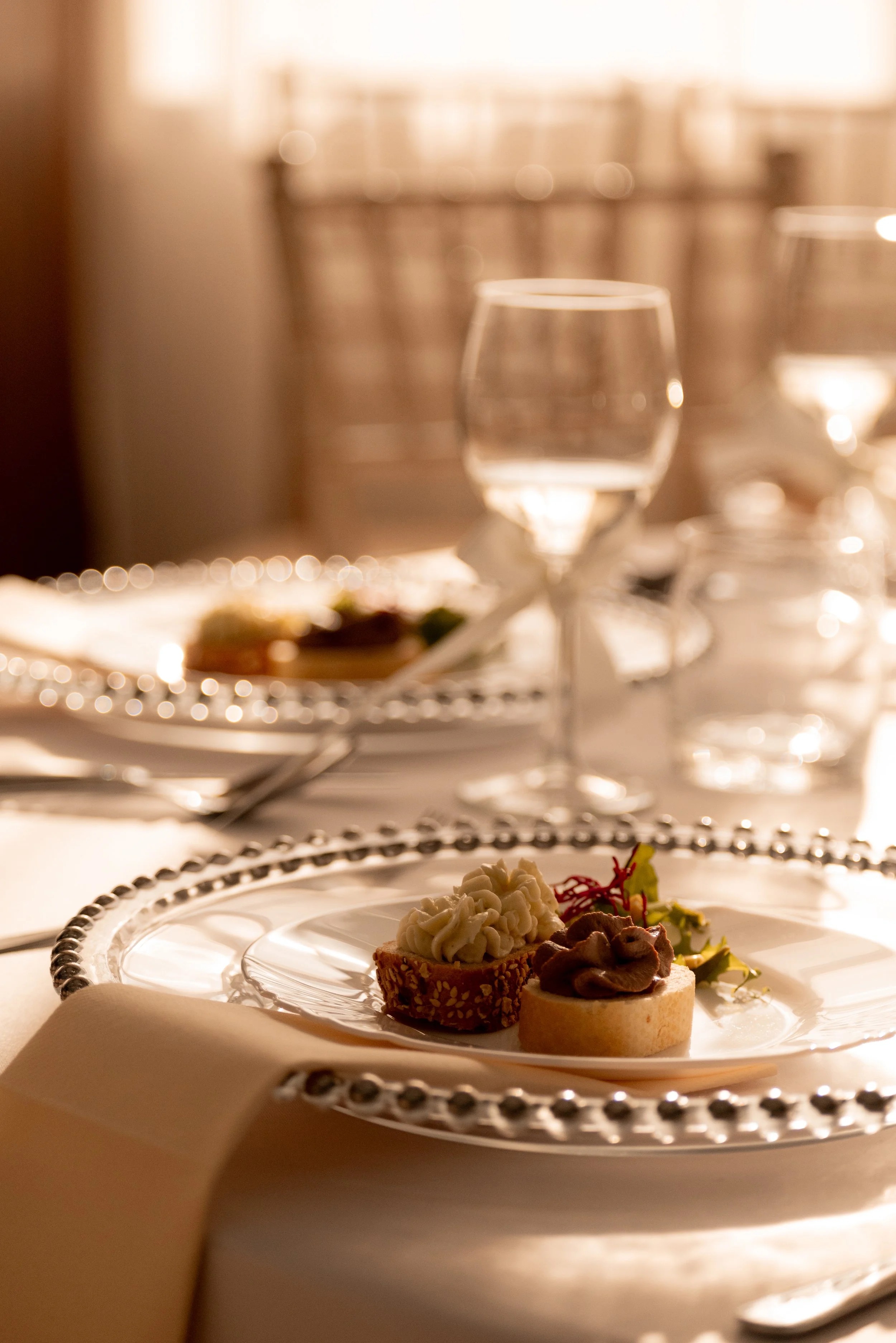Elegant table setting with a plate of two decorated small desserts, wine glasses, water glasses, and a napkin, in a warmly lit dining environment. Photographed by Andrea Cabajova, Girl with Bandana.