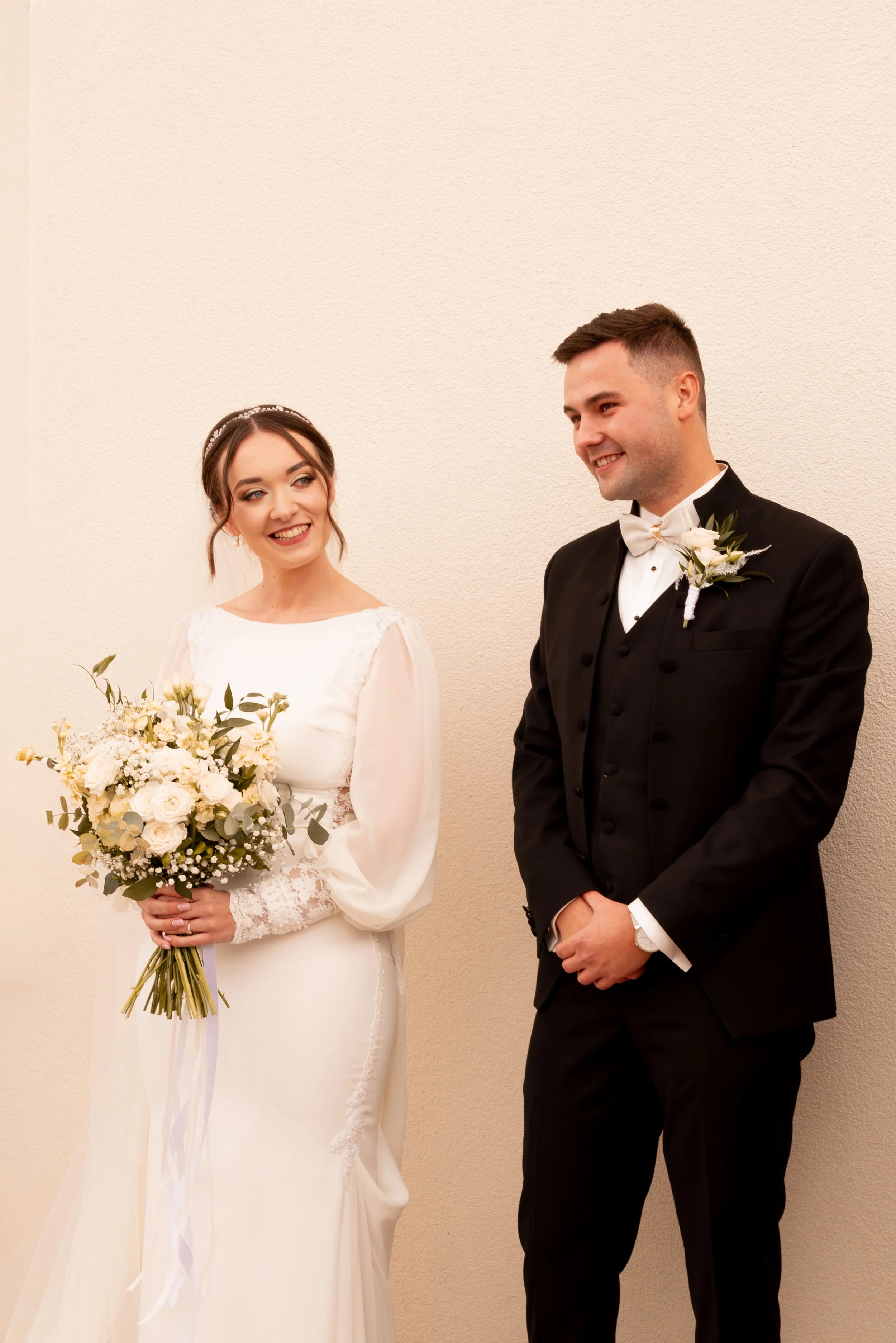 Bride and groom standing together indoors, bride holding a bouquet, both smiling and looking at each other, against a plain beige wall. Photographed by Andrea Cabajova, Girl with Bandana.