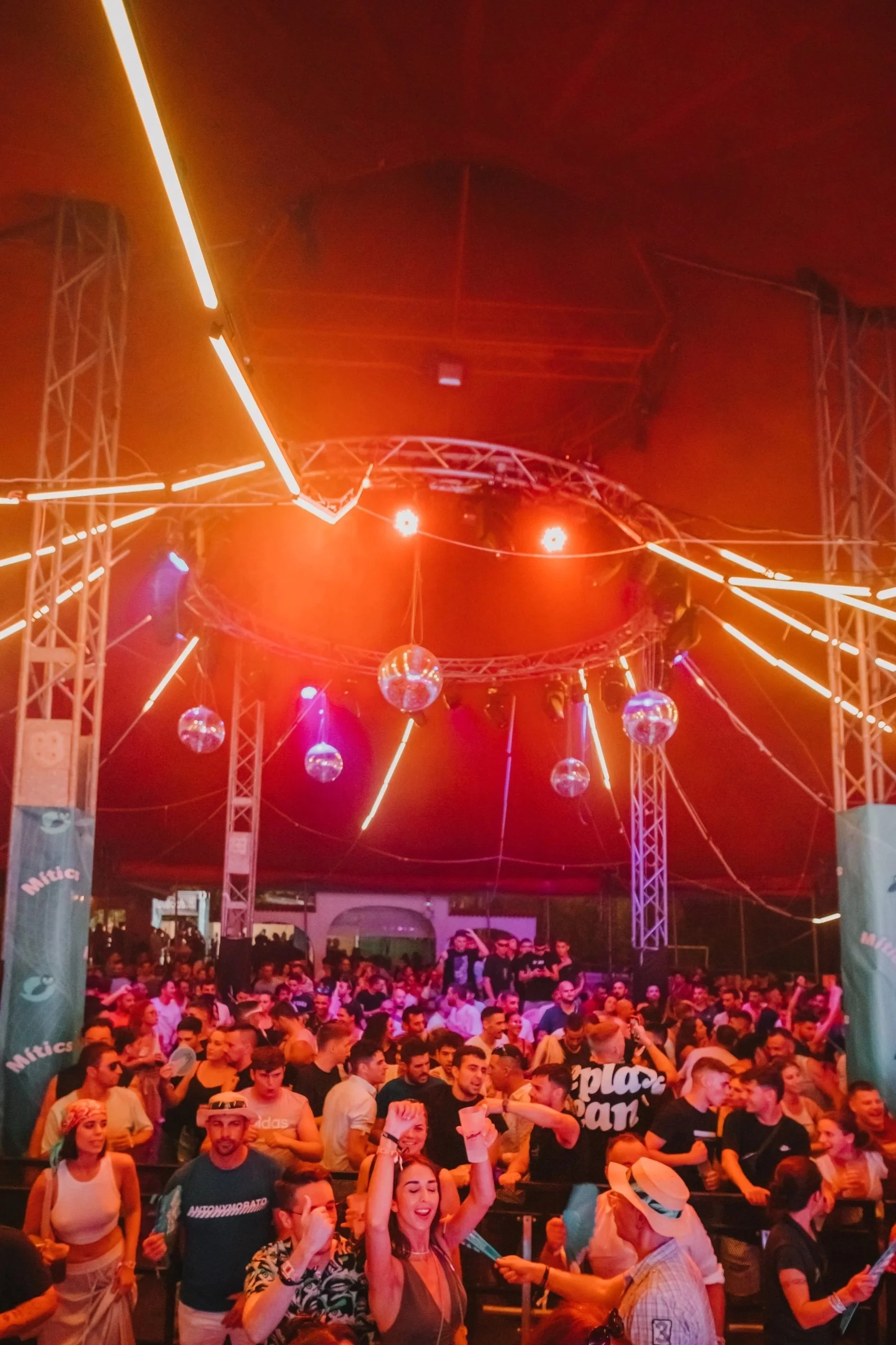 Crowd of people at a nightclub or concert with neon lights and disco balls overhead.