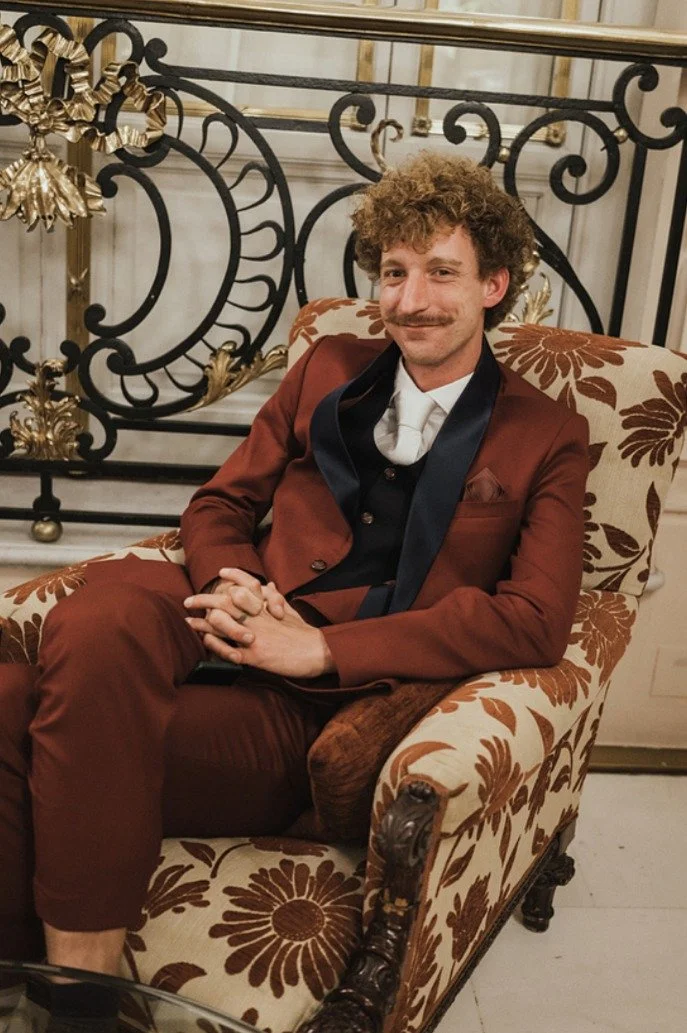 A man with curly hair and a mustache, dressed in a maroon suit with a white shirt and black tie, sitting comfortably in an ornate armchair with floral upholstery in a luxurious setting.