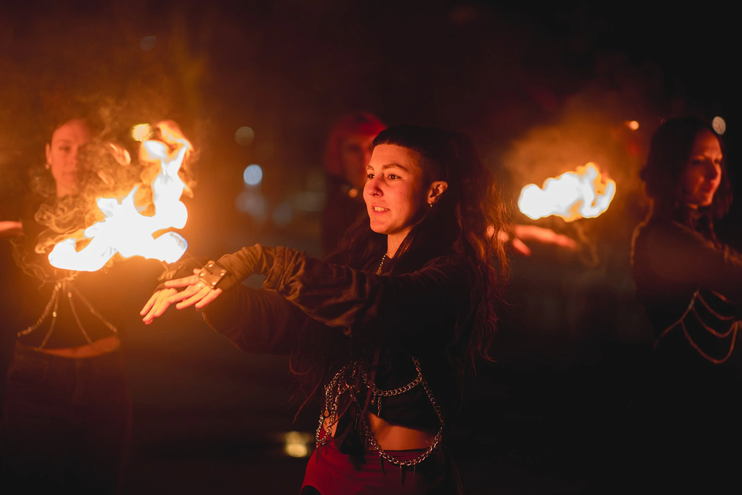 A group of women performing a fire dance at night, holding flames in their hands, with a dark background and their faces illuminated by the fire.