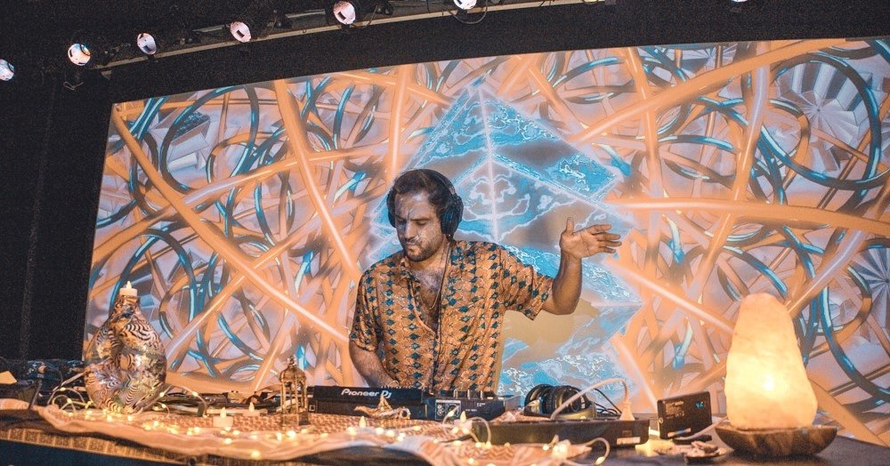 A DJ wearing a patterned shirt and headphones is performing at a music event, with a colorful digital backdrop and decorative lighting around the DJ booth.