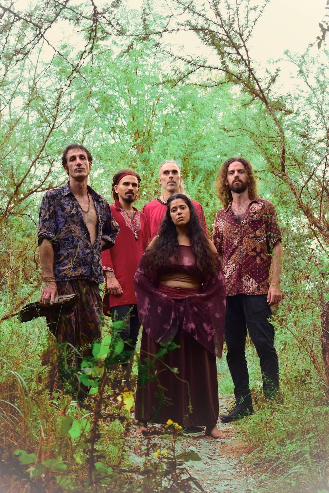 A group of five people standing in a forested area, surrounded by green trees and bushes, looking at the camera.