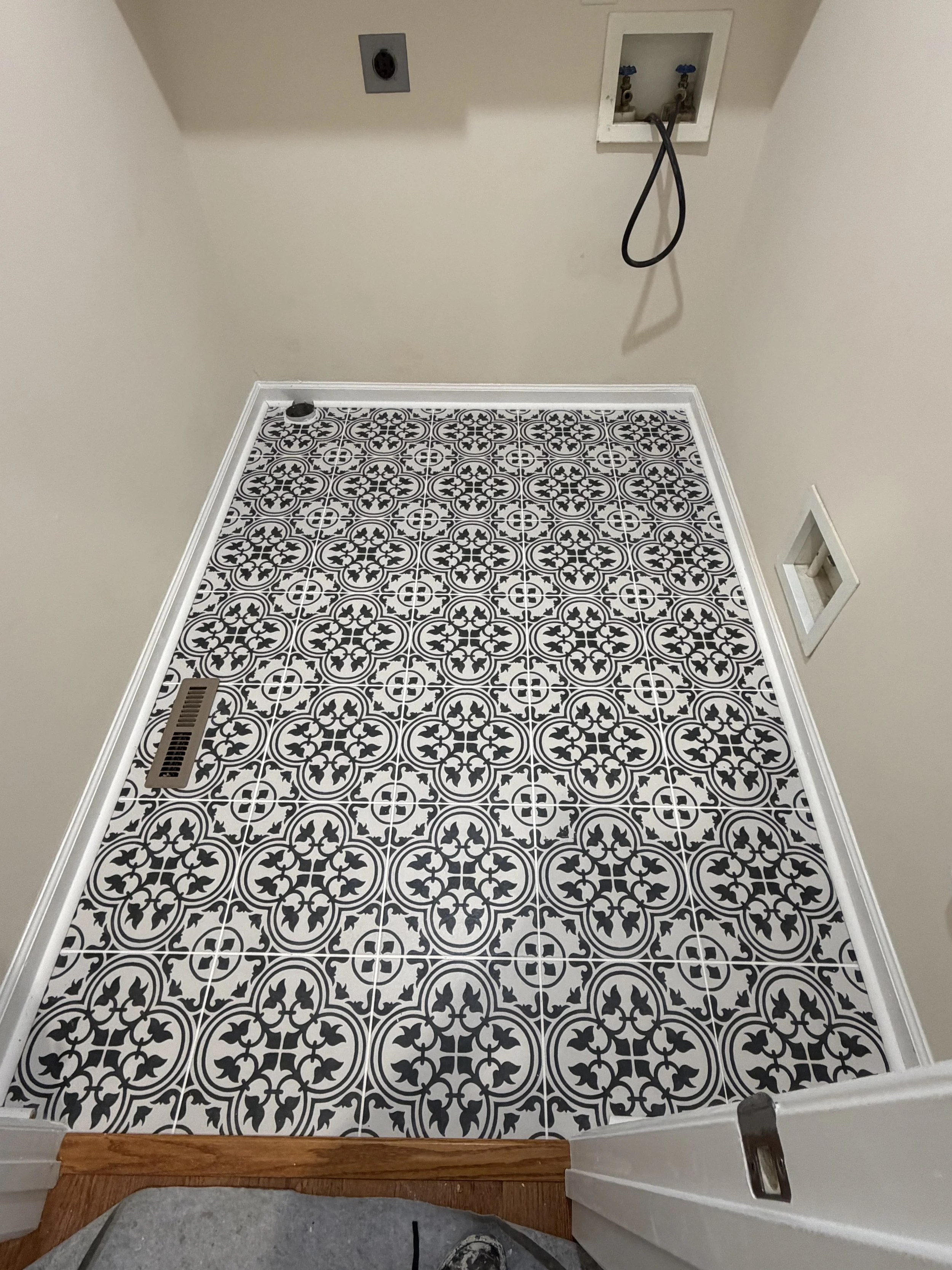 A laundry room floor with black and white patterned tiles, a vent, and an outlet near the floor. There is a wall with a plumbing hookup and an electrical outlet above it.