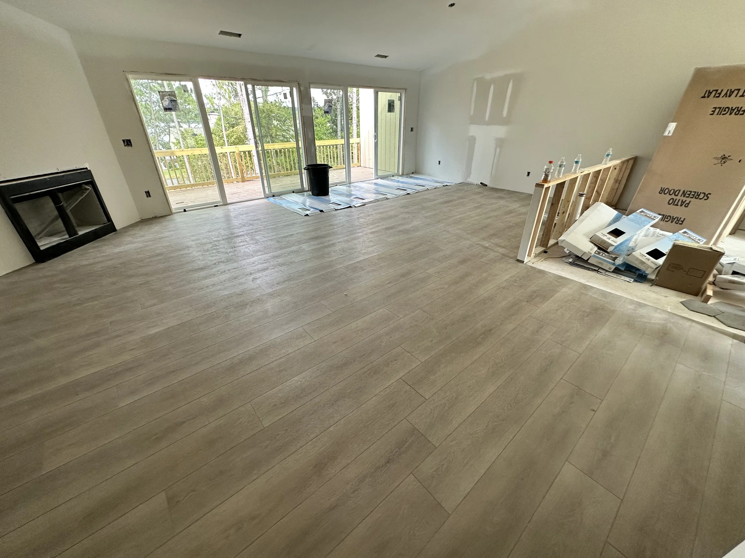 Interior of a room under construction with newly installed light wood flooring, a fireplace on the left wall, glass sliding doors leading to an outdoor deck with a yellow railing, and construction materials and tools scattered around.